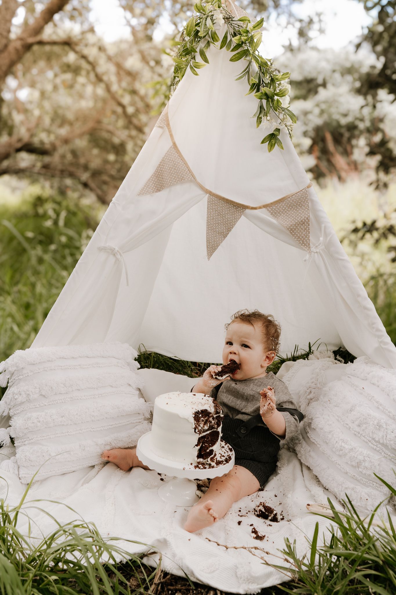Outdoor cake smash session with a baby seated in front of a teepee, surrounded by long grass. A whimsical, natural setting with playful, mes
