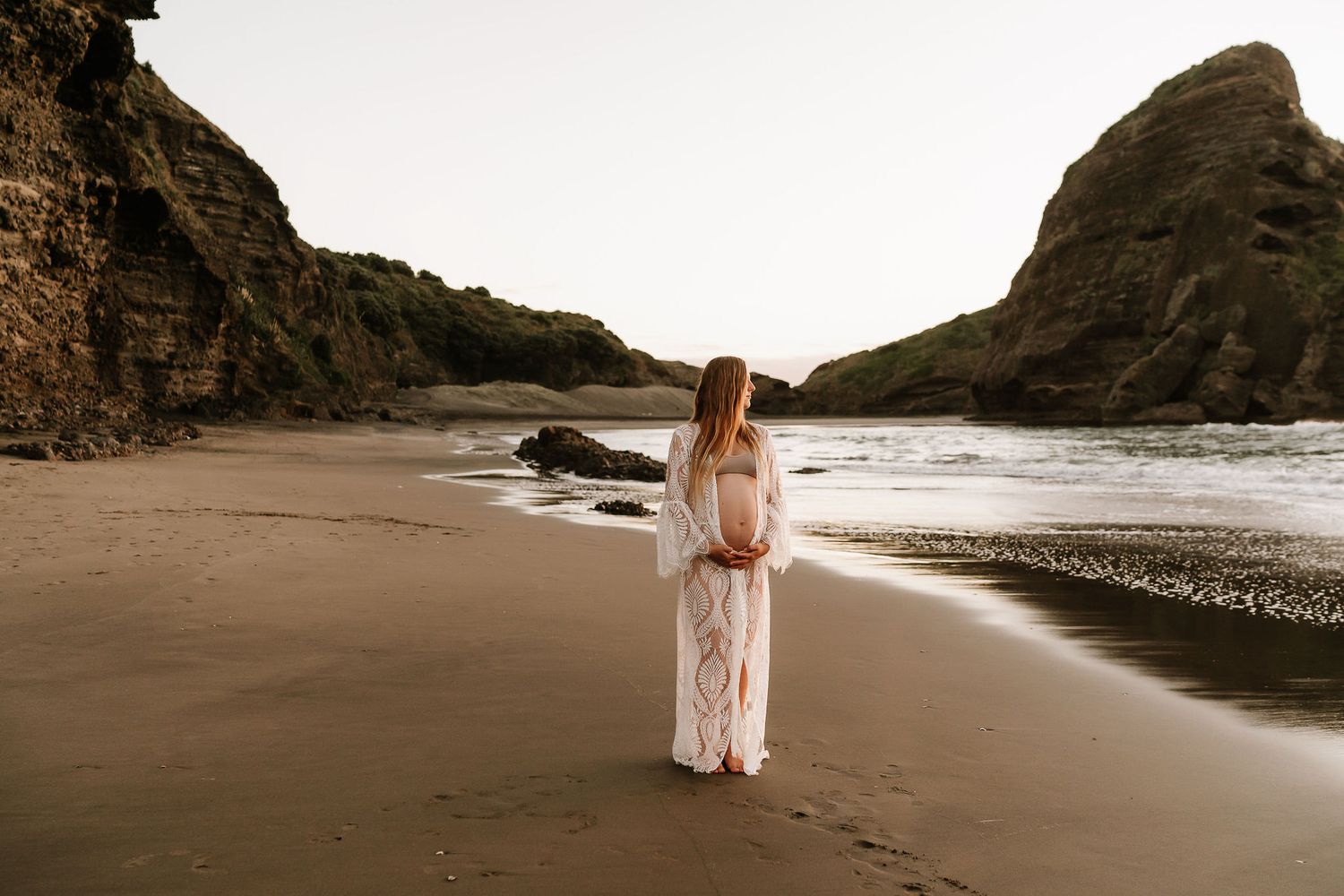 woman is wearing a white dress and is holding her baby belly as the sun sets behind her on the beach