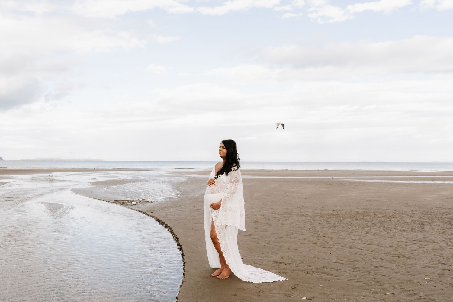 woman is wearing a white dress and is holding her baby belly as the sun sets behind her on the beach