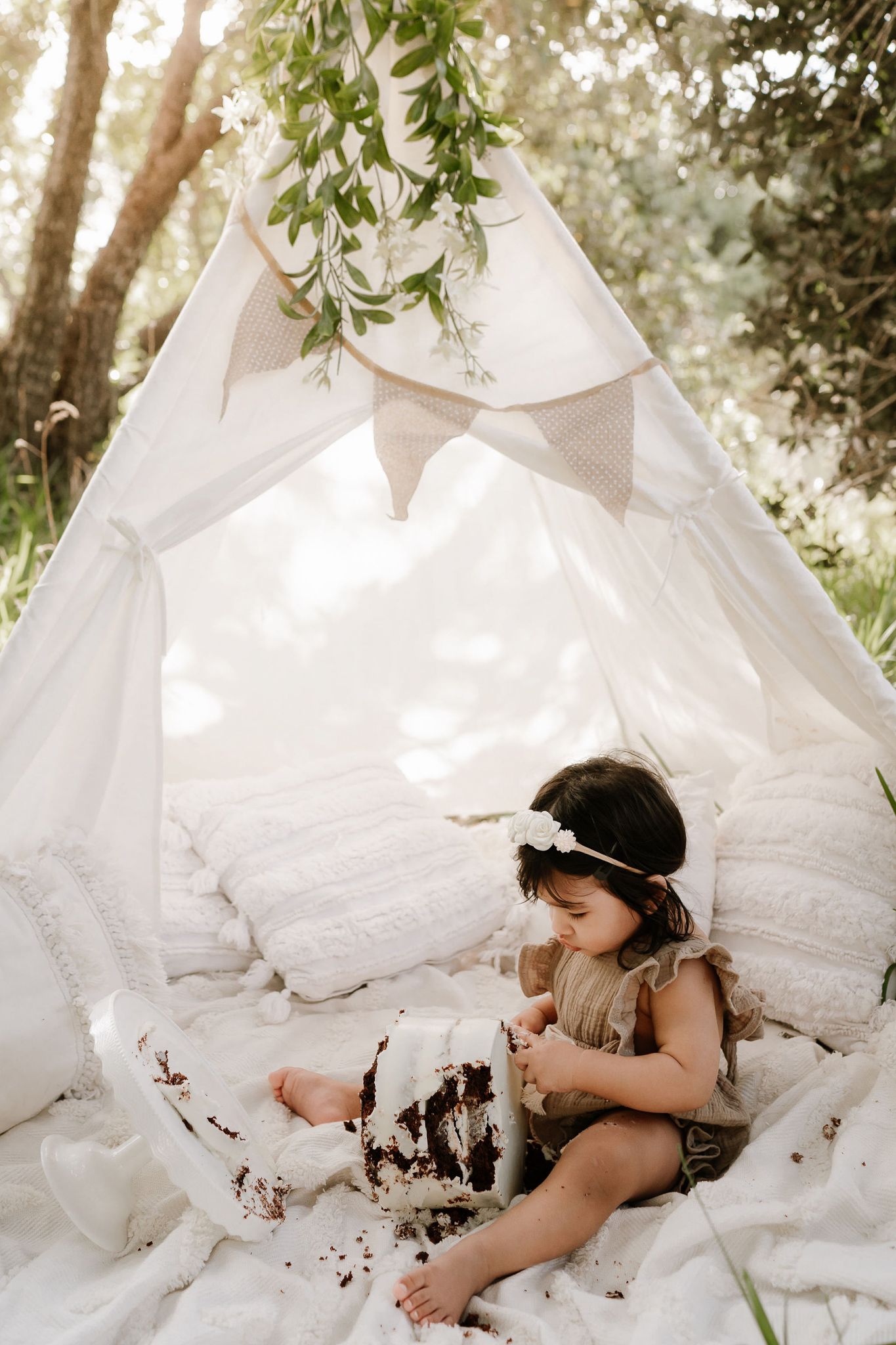 Outdoor cake smash session with a baby seated in front of a teepee, surrounded by long grass. A whimsical, natural setting with playful, mes