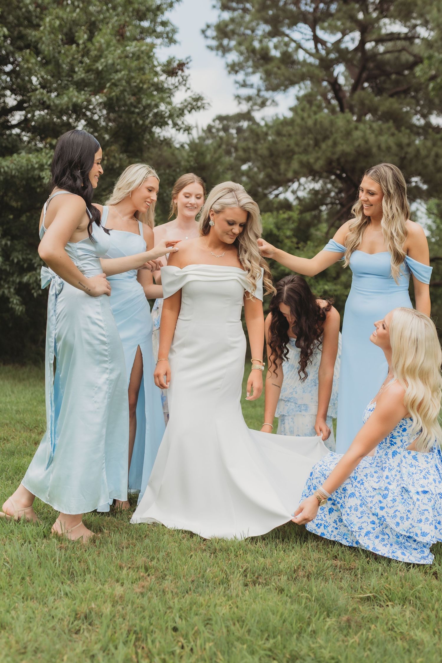 A bridal party in light blue bridesmaid dresses and a traditional white wedding gown stand together on grass outdoors.