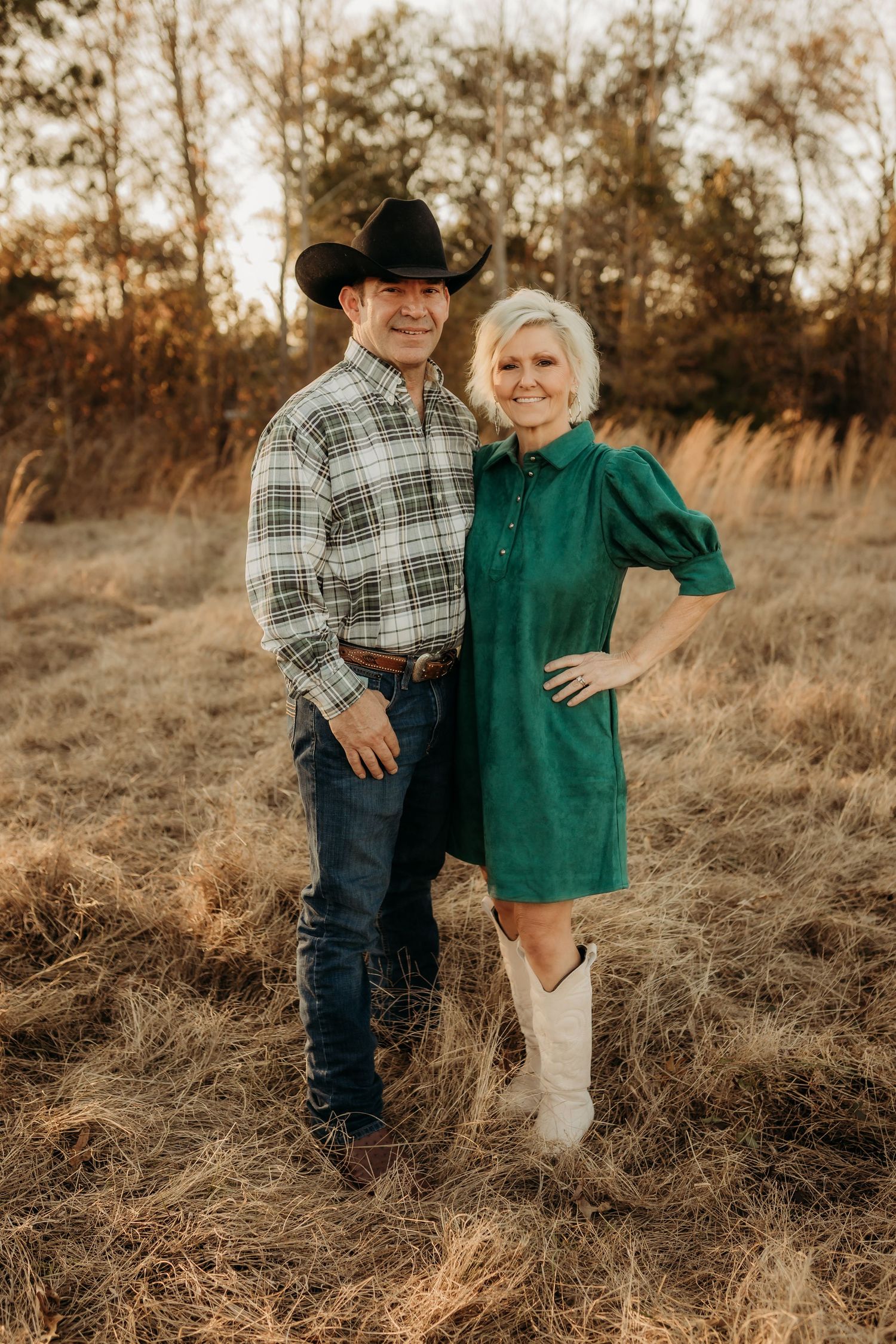 A couple in western attire embraces in a golden field at sunset, creating a warm and romantic outdoor portrait.