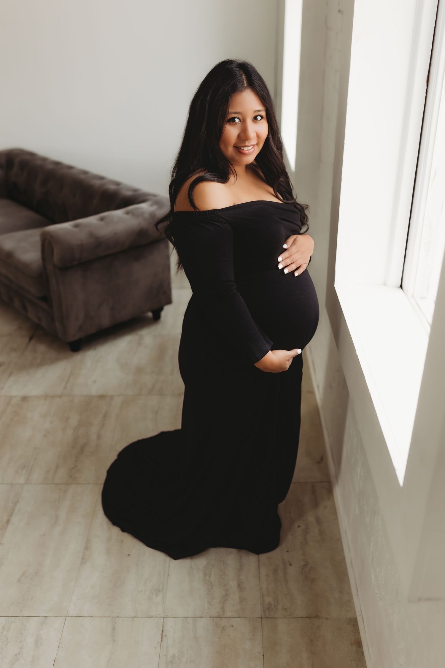 Expectant mother in black formal gown poses by window during maternity photoshoot.