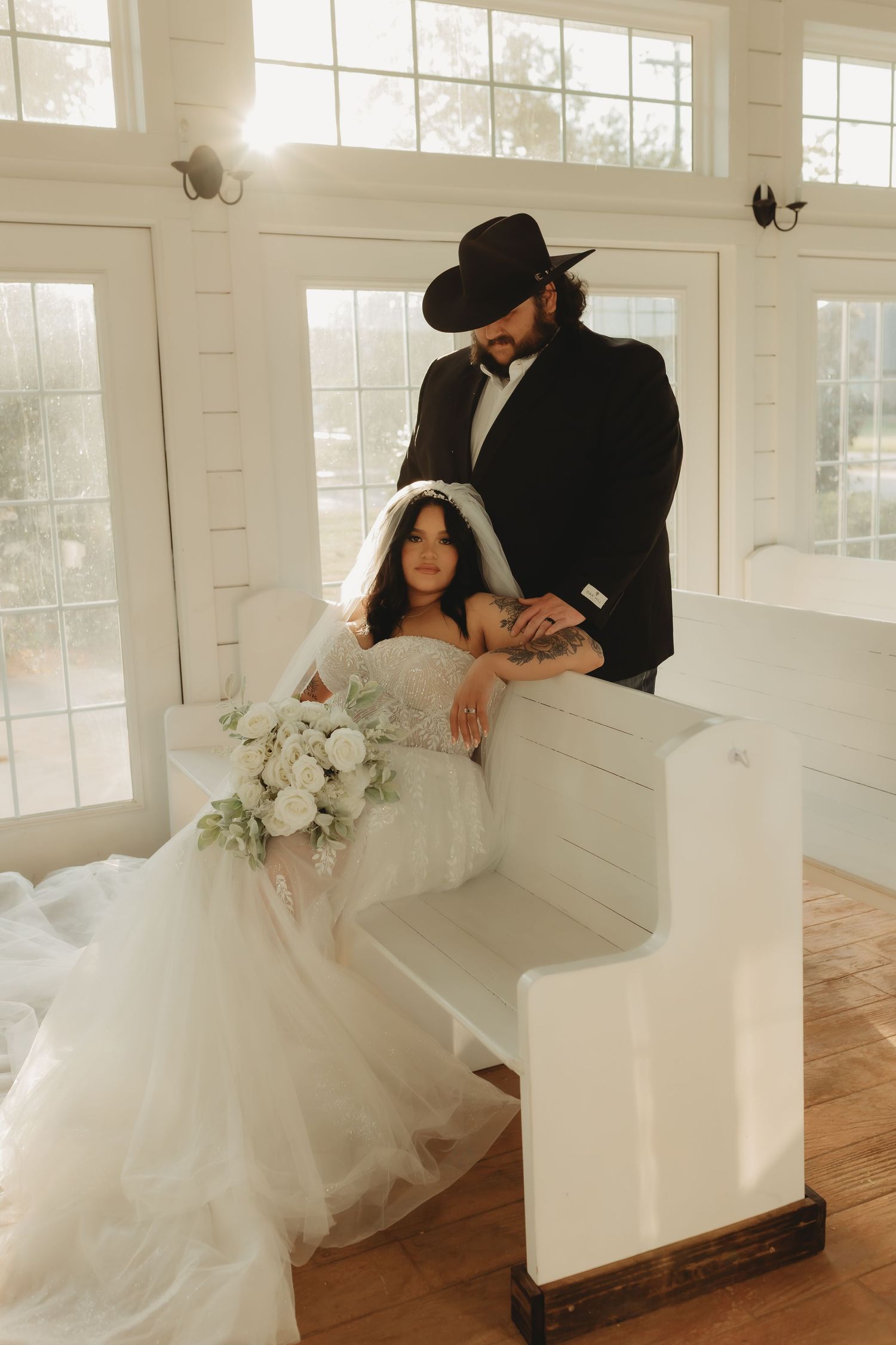 A romantic wedding photo in a bright white chapel with a bride in a flowing gown and groom in black western attire.