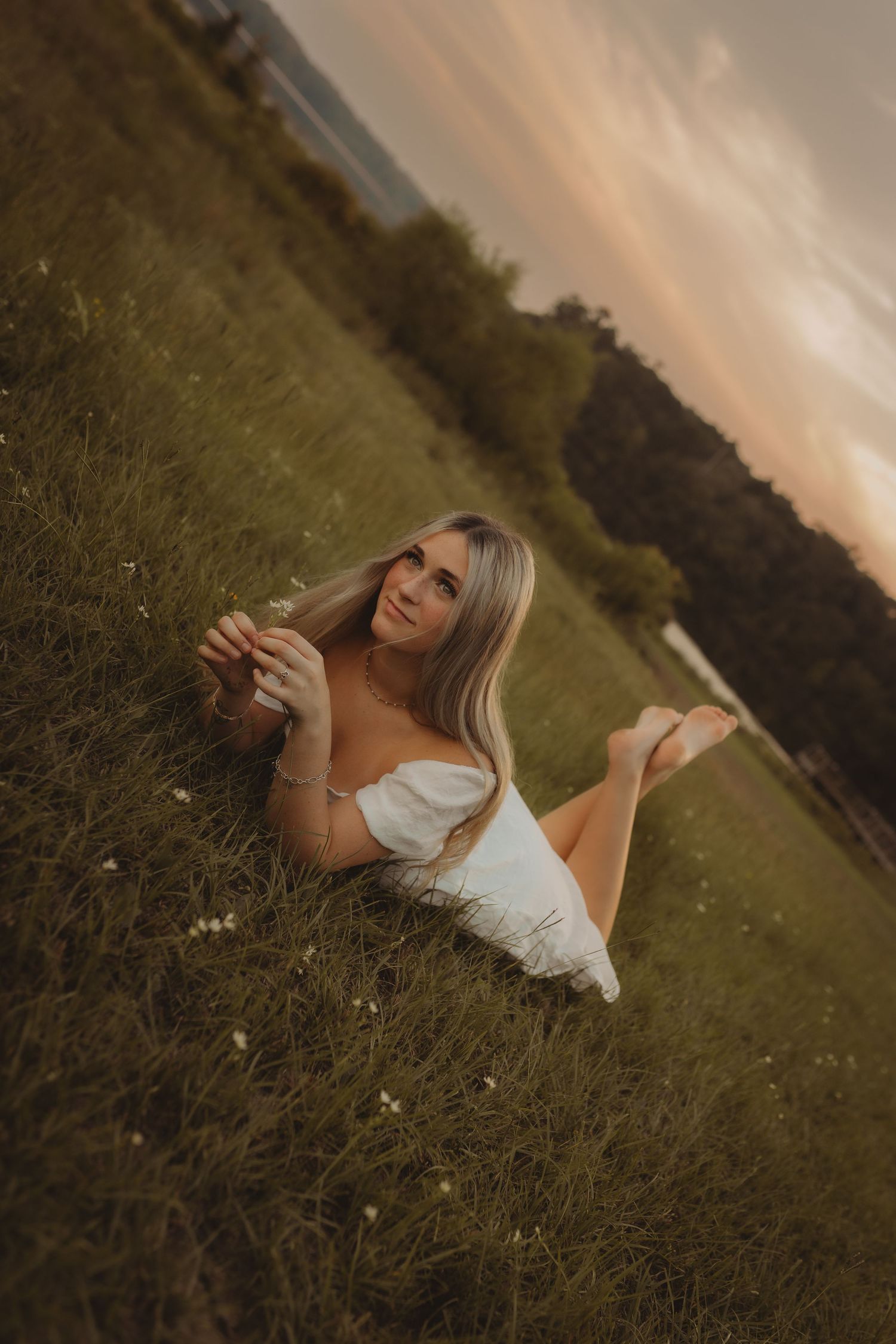 A woman in a white dress lies on grassy ground during sunset, creating a dreamy outdoor photoshoot atmosphere.