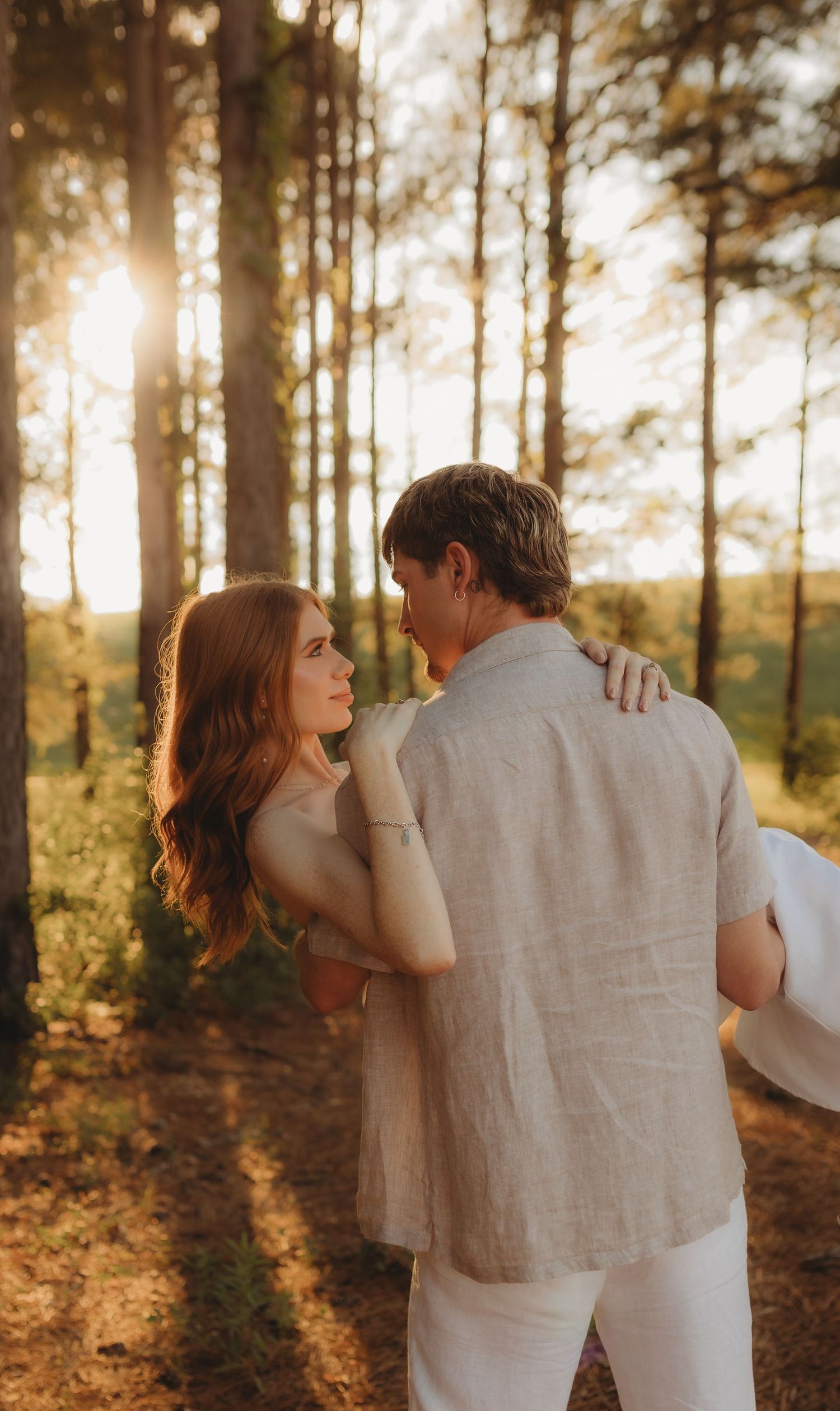 A couple in light-colored clothing embraces while walking through a sunlit forest during golden hour.