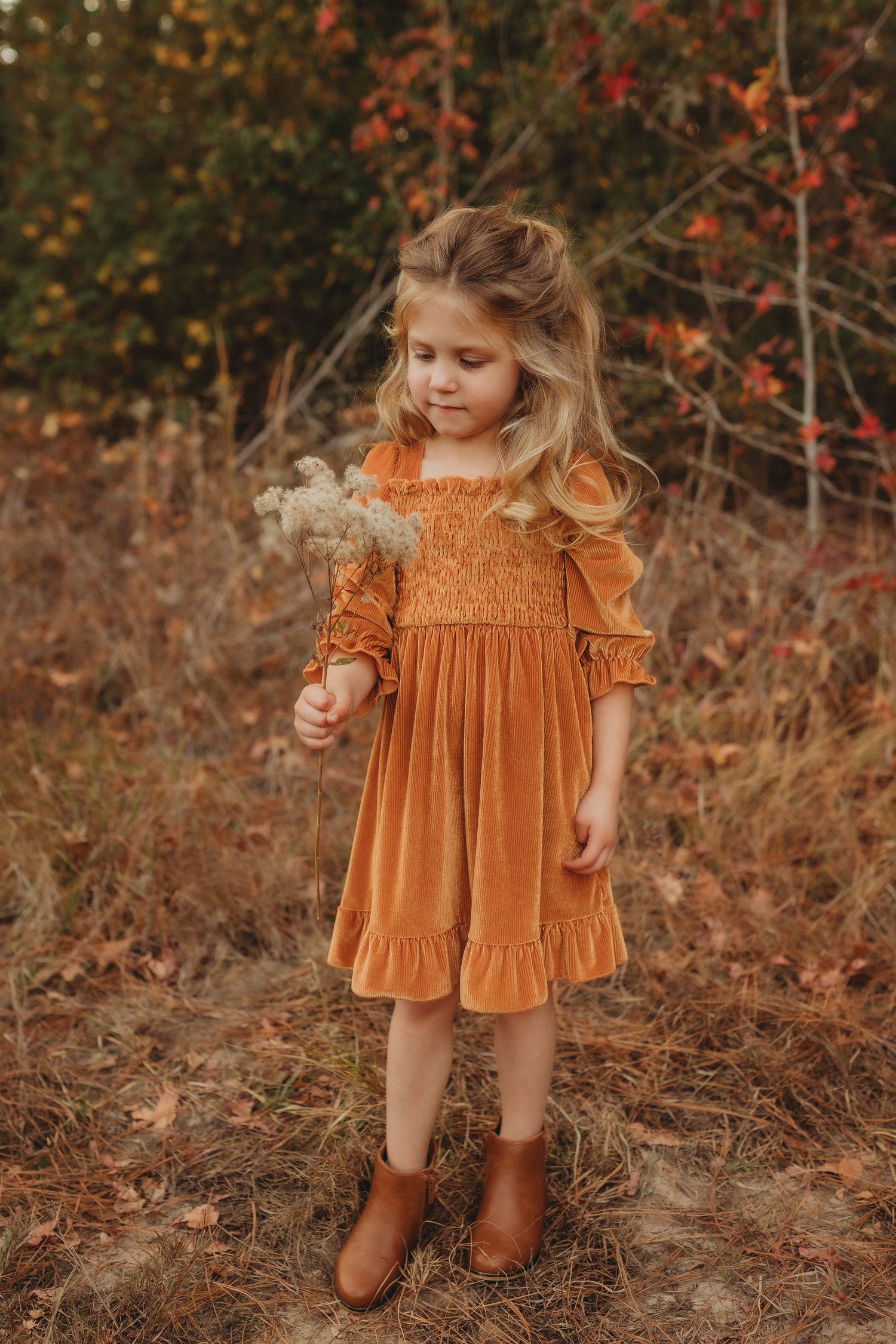 A child in a rust-colored velvet dress stands among autumn foliage with brown boots.