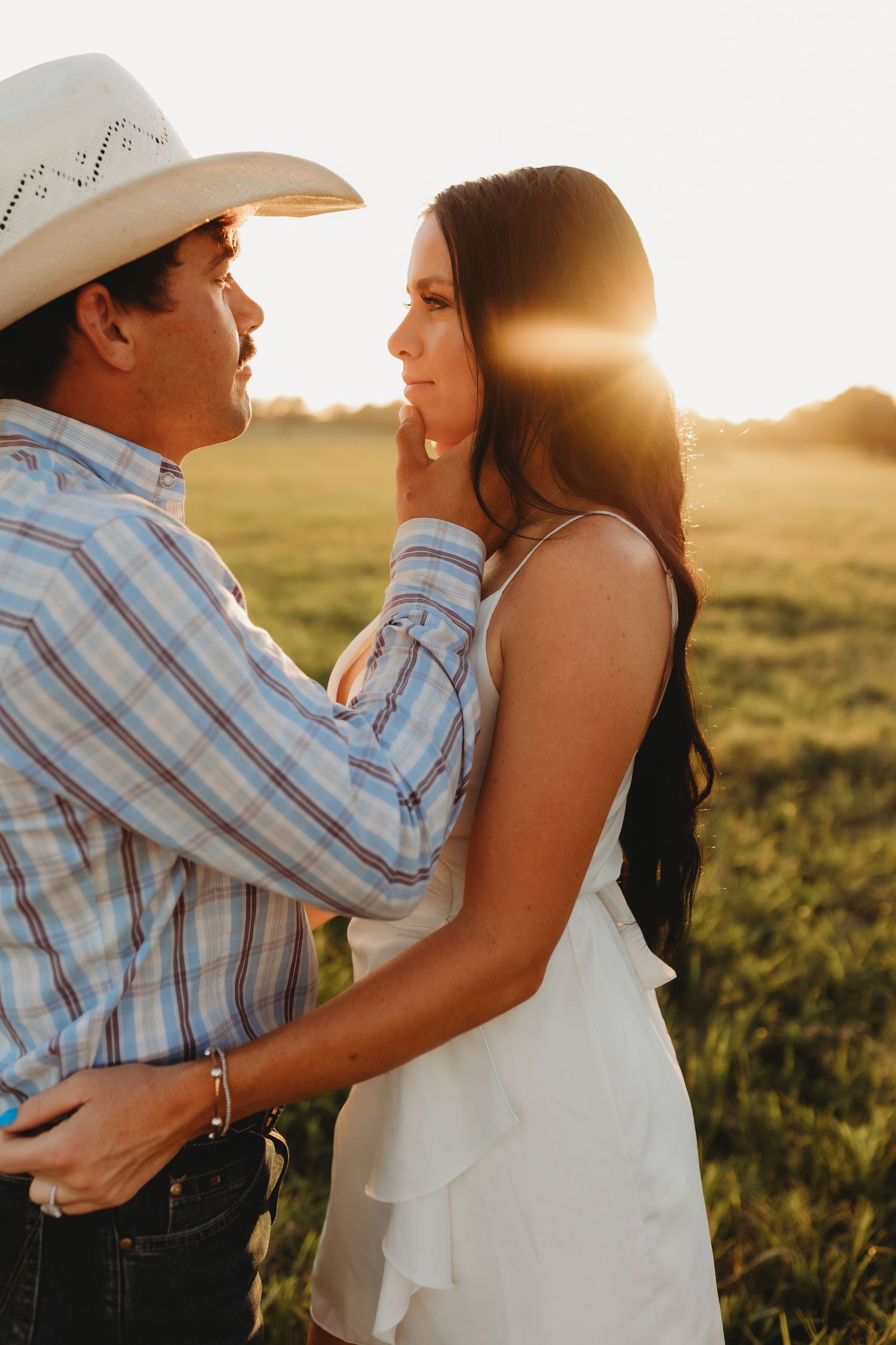 A romantic couple embraces in a field at sunset, with sunlight glowing behind them as they share an intimate moment.