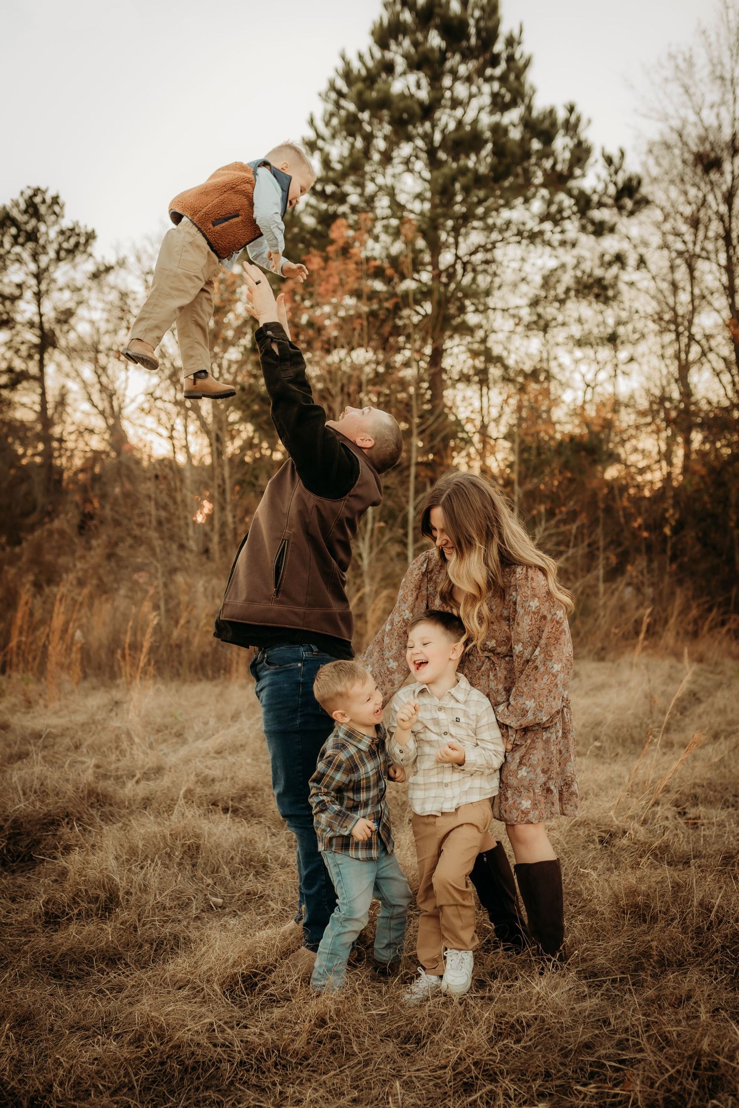 A family in casual attire playfully jumps together in a golden field at sunset while surrounded by pine trees.