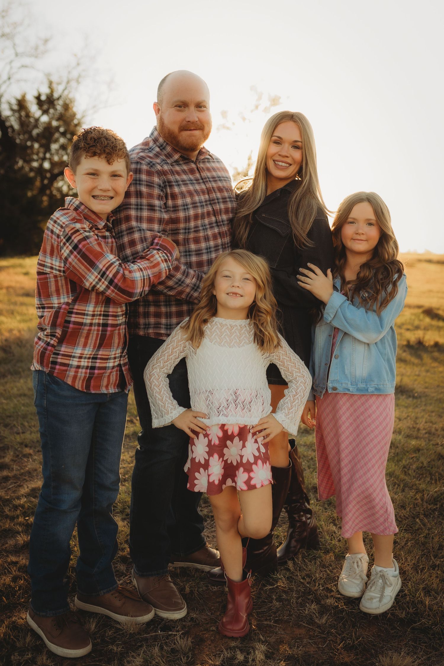 A family poses together outdoors at sunset wearing coordinating plaid and casual outfits in warm autumn colors.