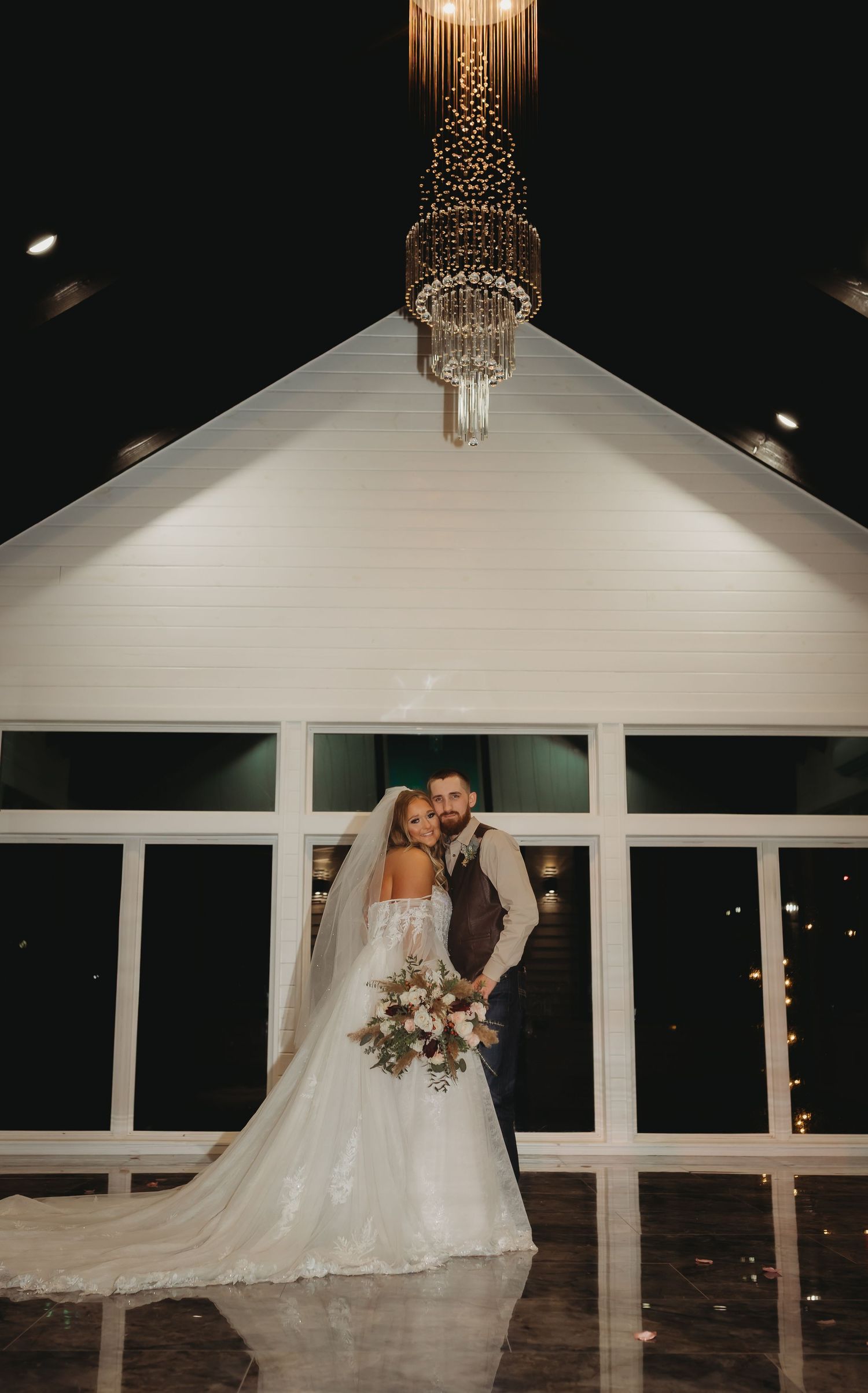 Elegant wedding venue interior features vaulted ceiling with crystal chandelier and large windows at night.