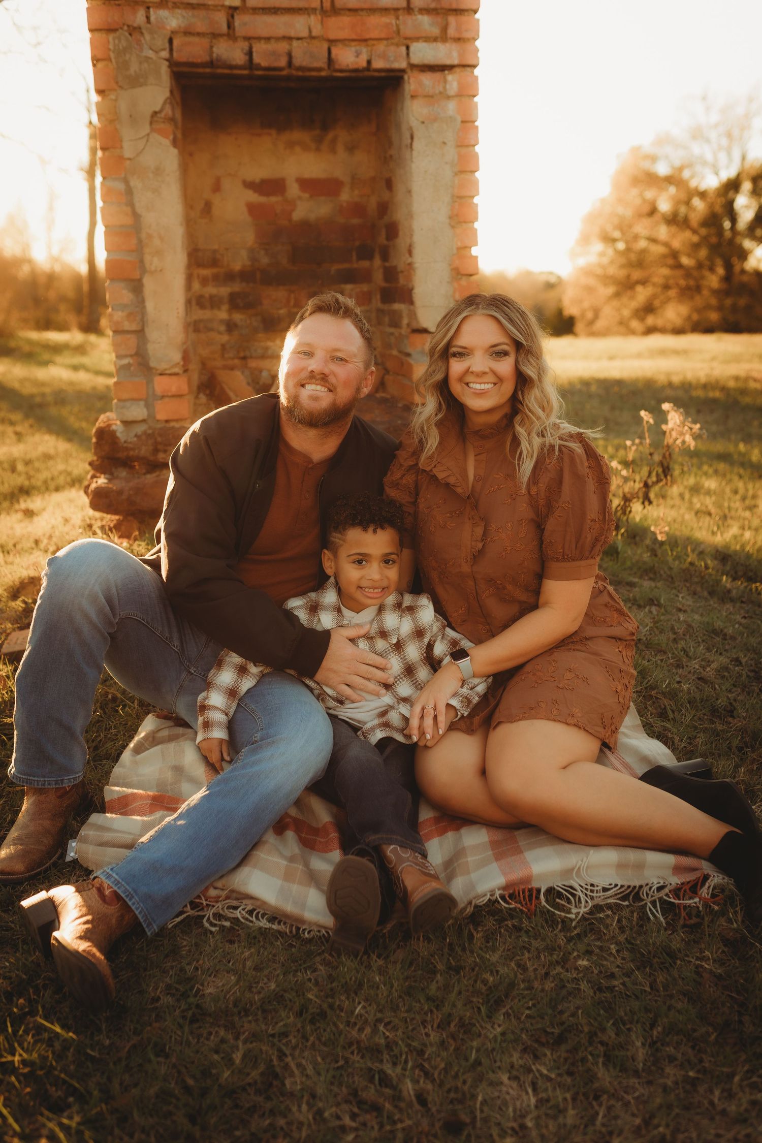 A family sits together on grass near a brick wall during golden hour sunset, sharing intimate moments in casual attire.