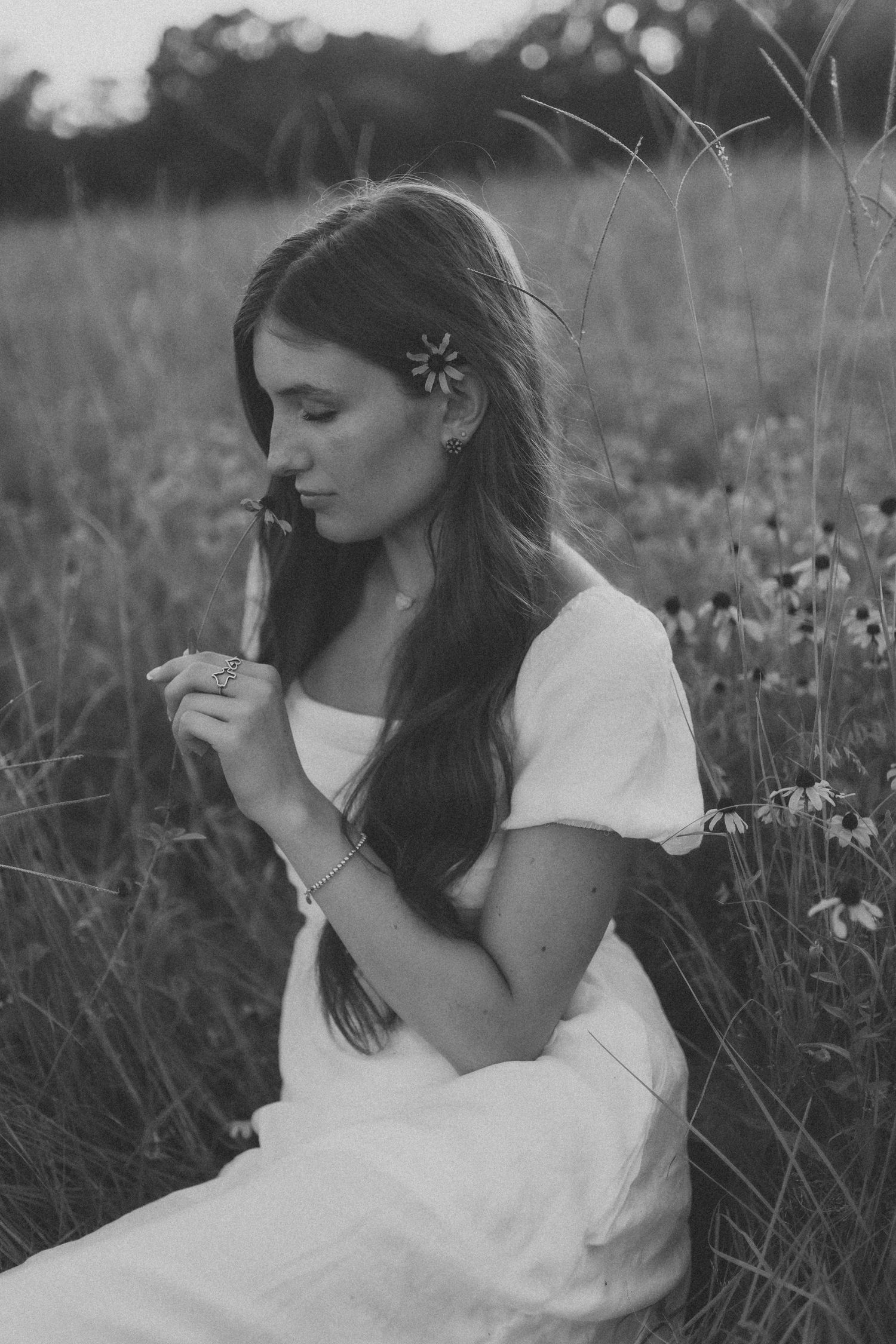 A serene black and white photo of someone in a white dress sitting in a wildflower meadow with a flower in their hair.