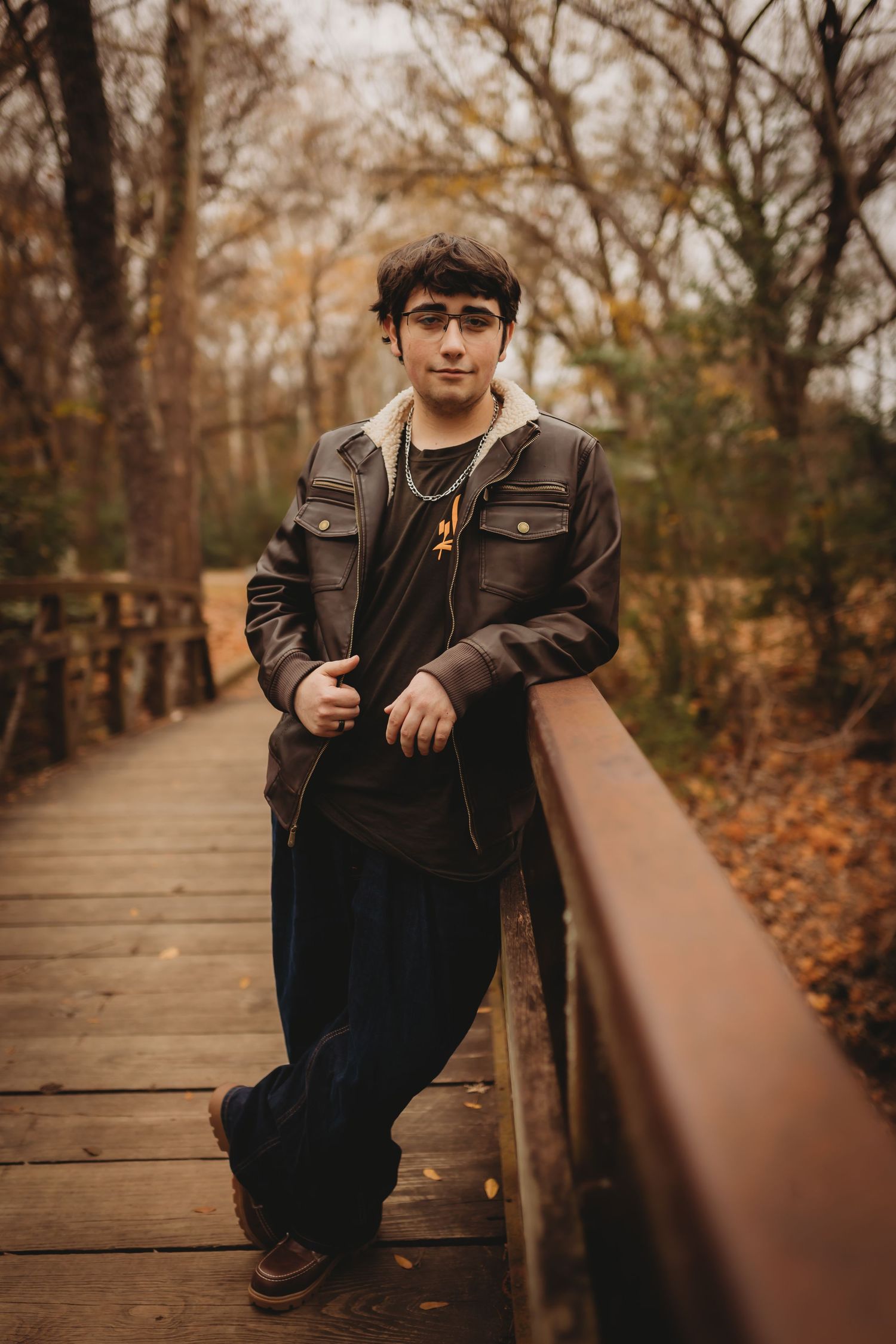 A person in a dark jacket sits on a wooden boardwalk surrounded by autumn trees and fallen leaves.