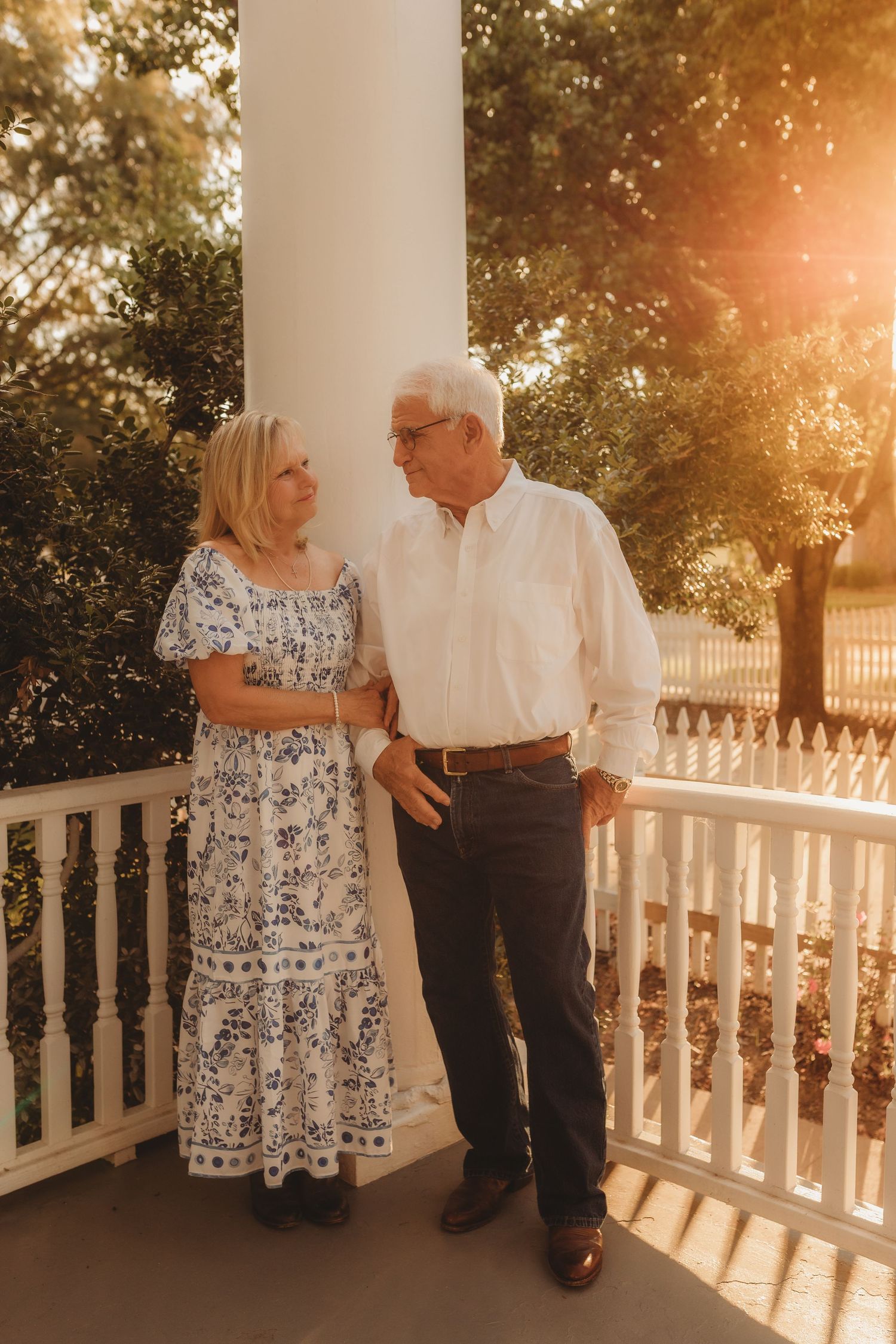 Elderly couple shares tender moments on a sunlit porch with white railings during golden hour.