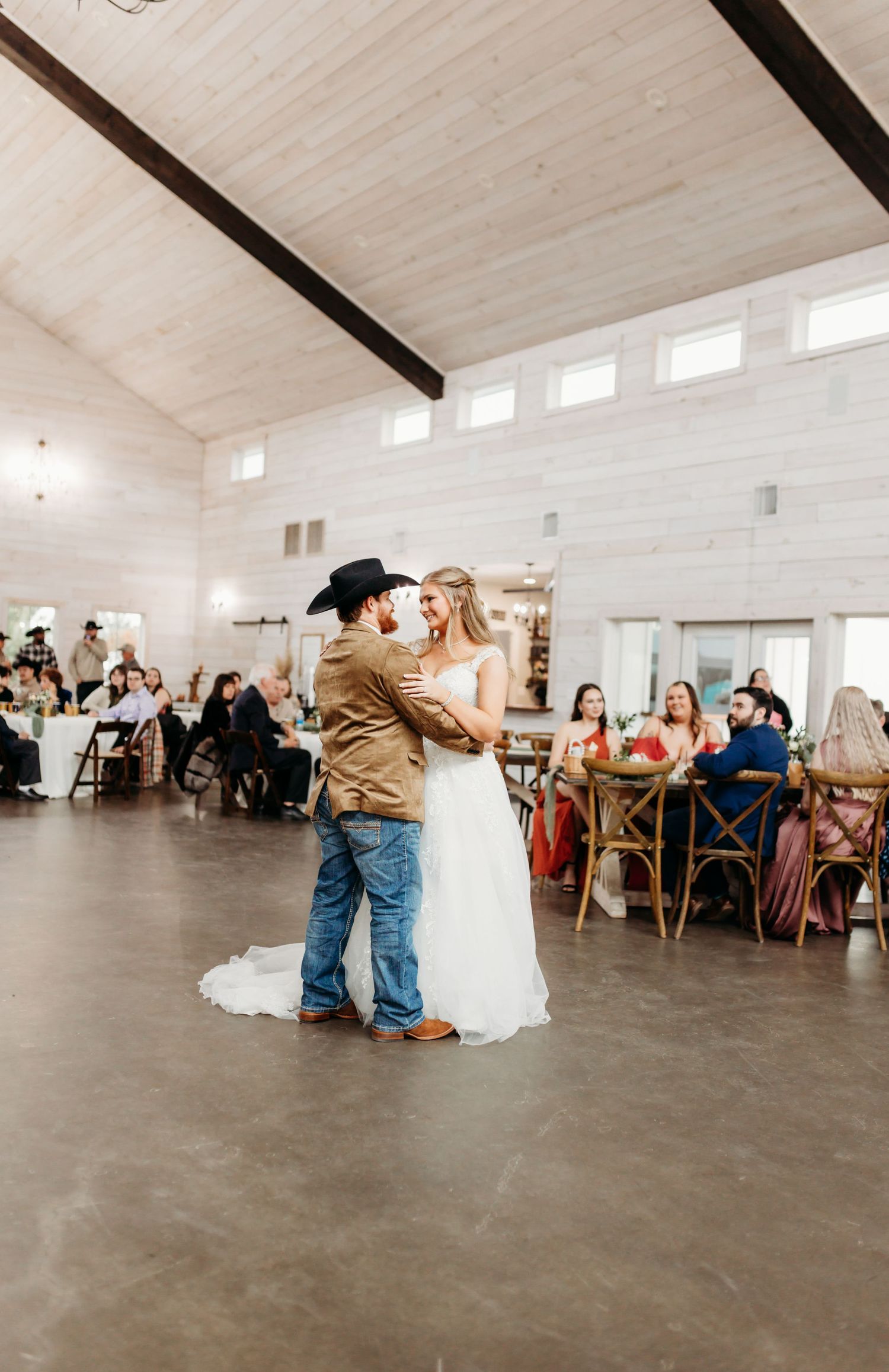A couple shares their first dance at a rustic barn wedding reception with guests seated at round tables.