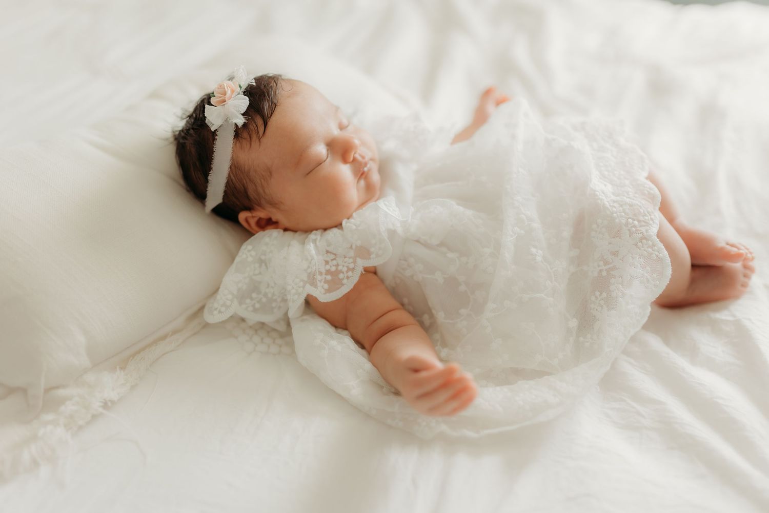 A sleeping baby wearing a delicate white lace dress and headband lies on soft white bedding.