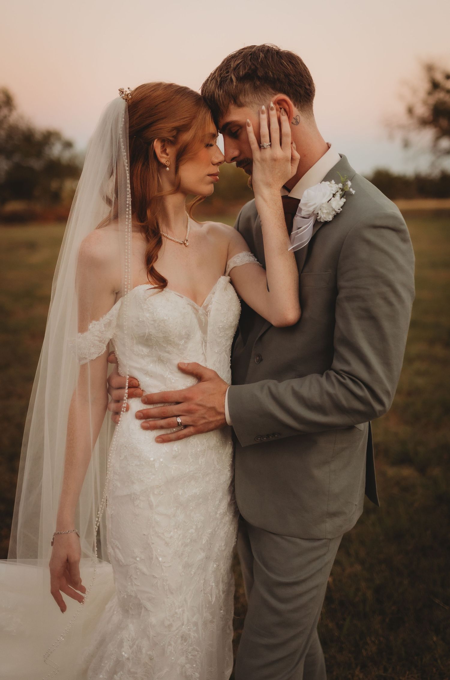 A romantic wedding couple embraces in a field at sunset, with the bride wearing a lace gown and flowing veil.