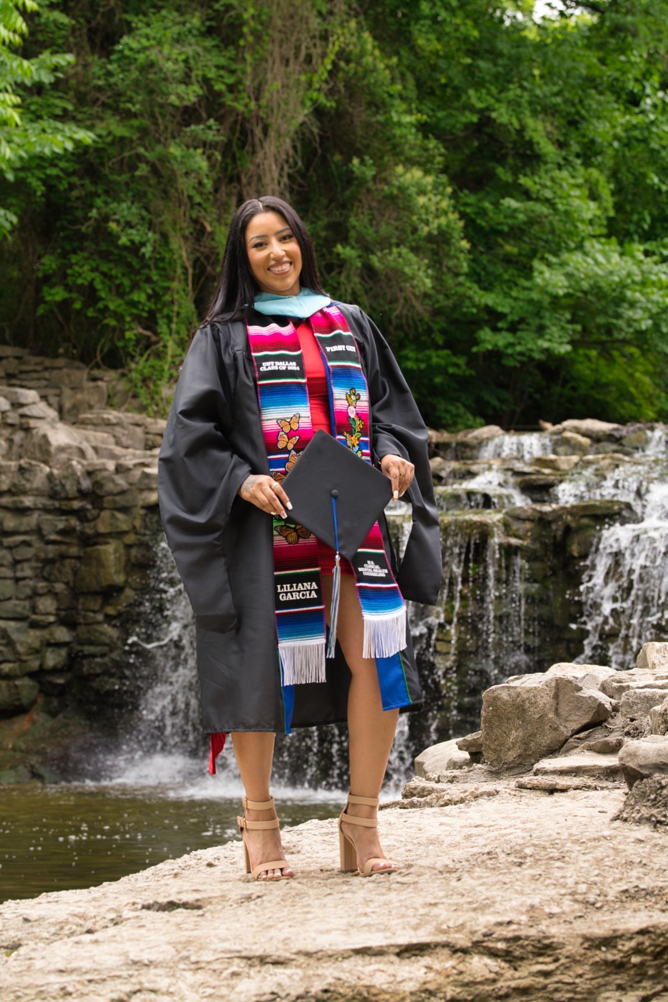 Young woman named Liliana Garcia in graduation gown and colorful stole poses by a waterfall holding her cap.