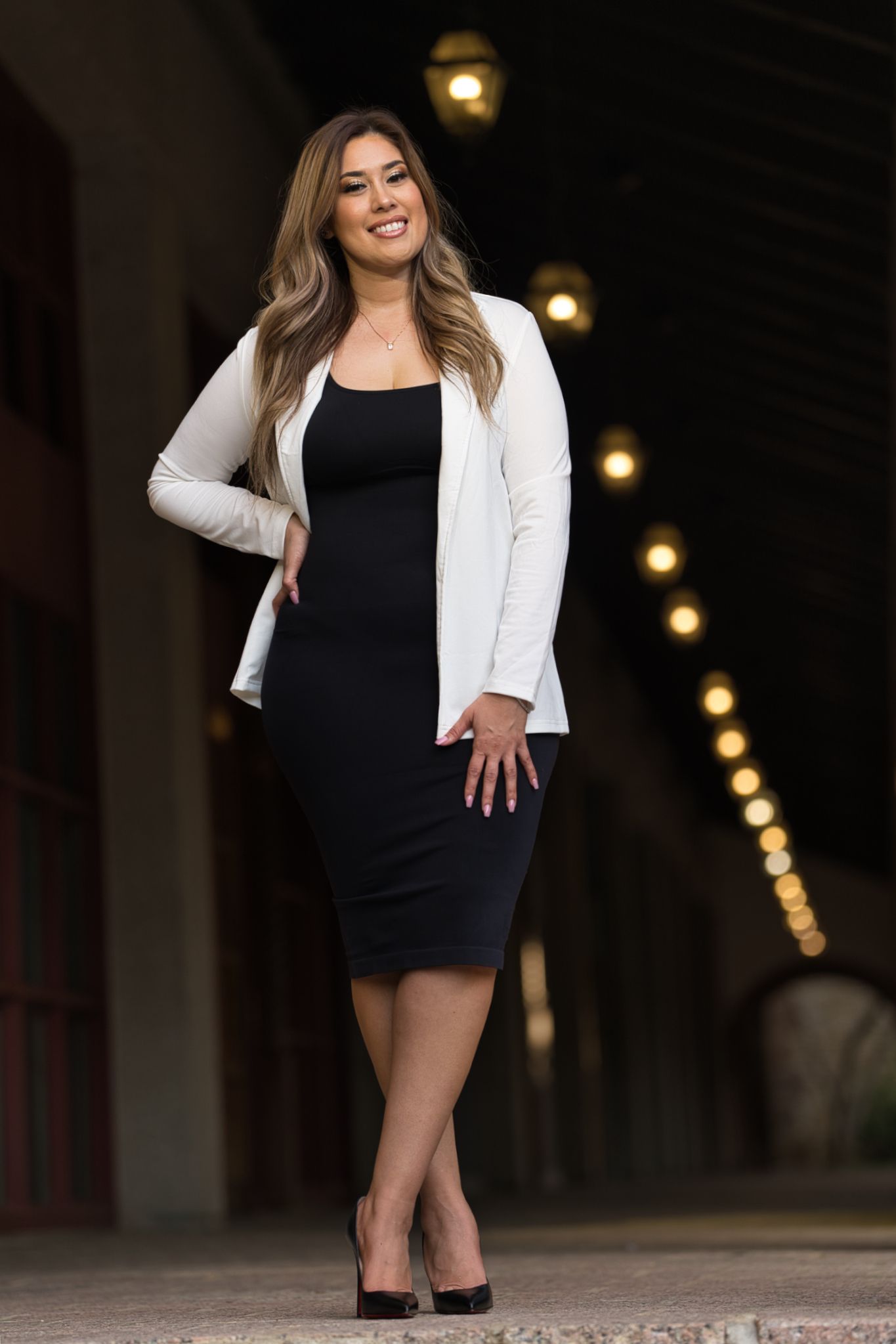 Woman in black dress and white blazer poses confidently in hallway with string lights.
