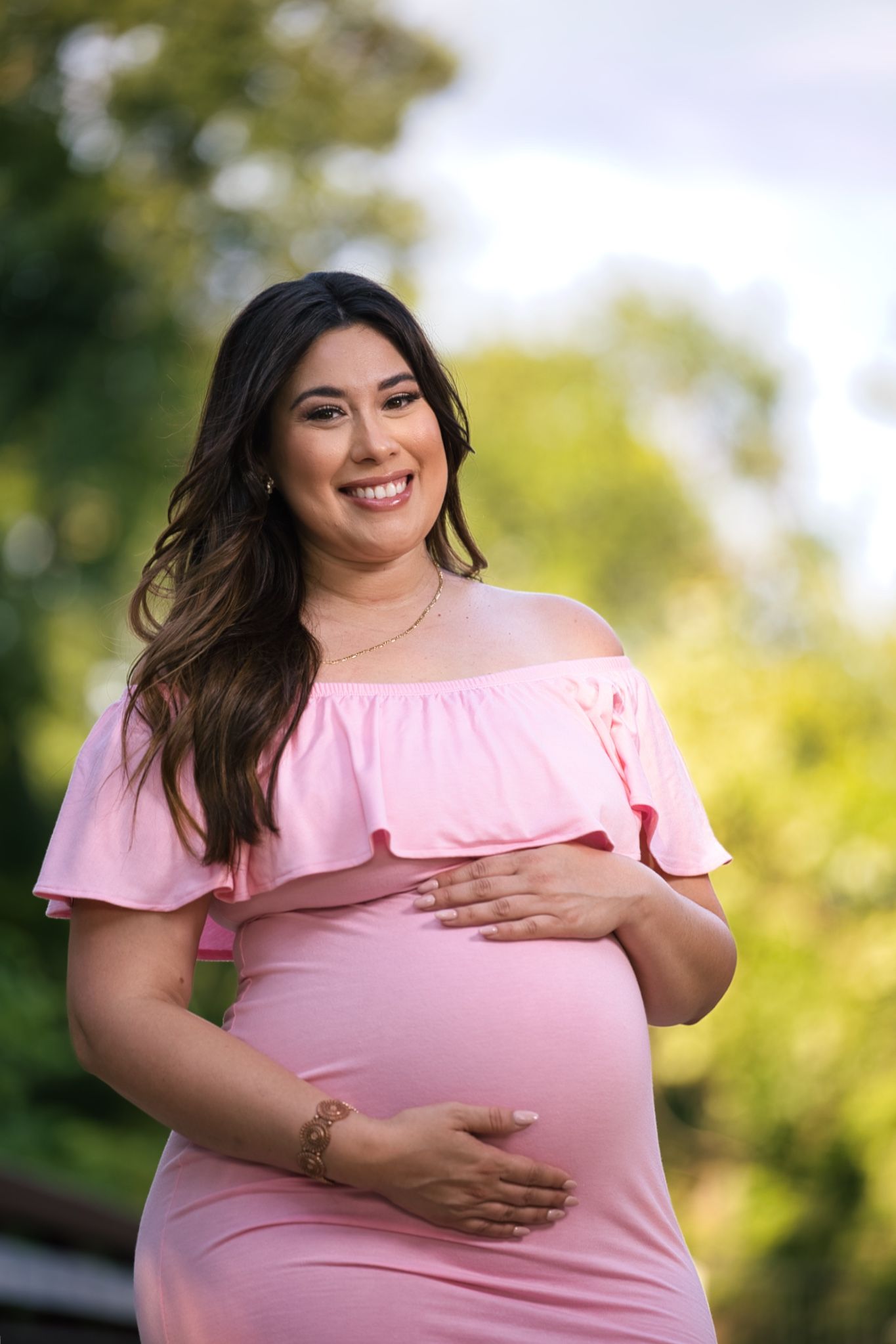 Glowing mother-to-be poses in pink off-shoulder dress during outdoor maternity session.
