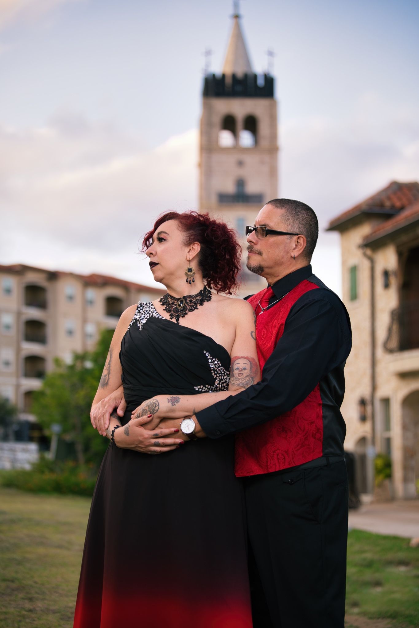 A couple in formal attire poses romantically in front of a church tower at dusk.
