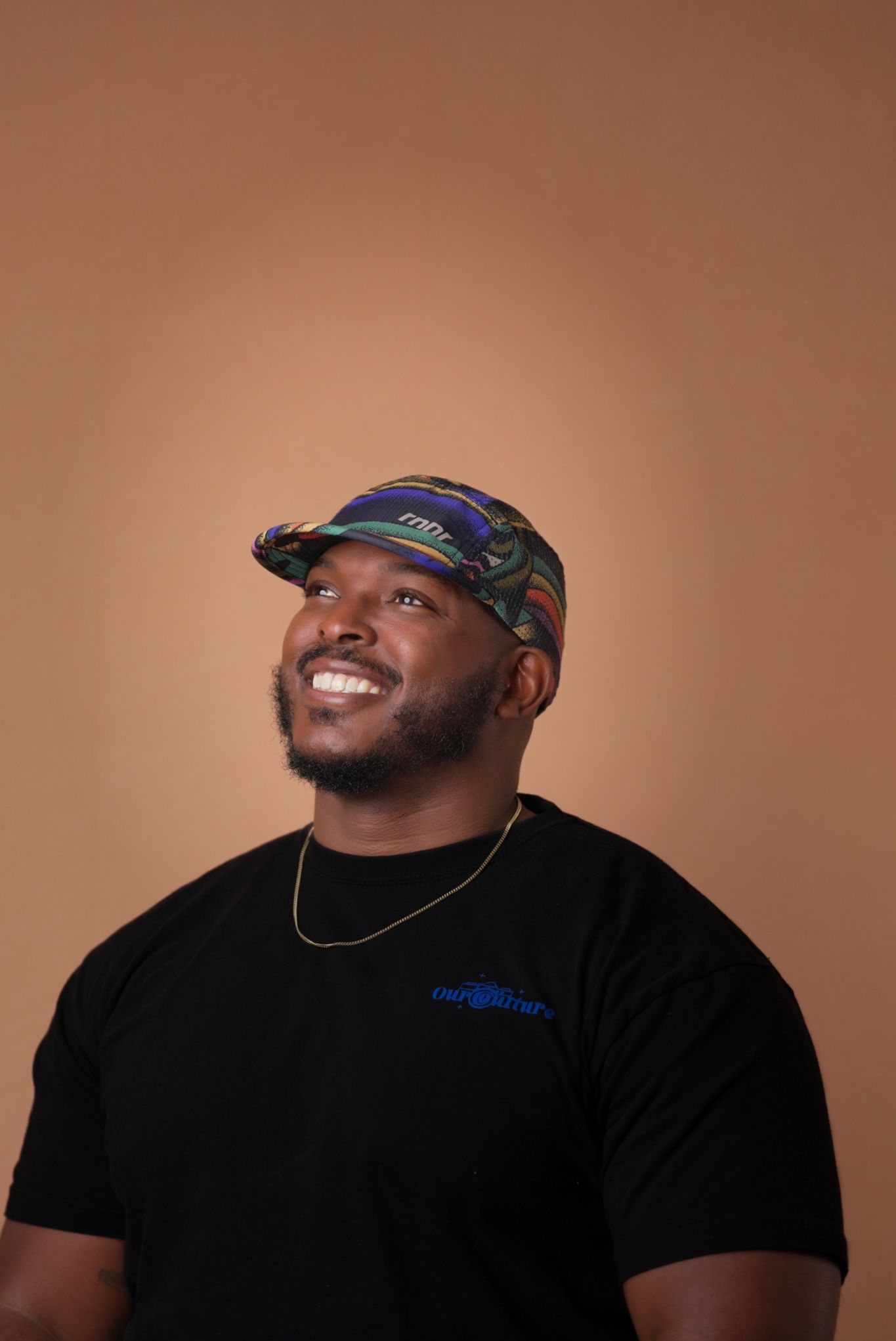 A person wearing a camouflage cap and black t-shirt smiles while looking up against a brown background.