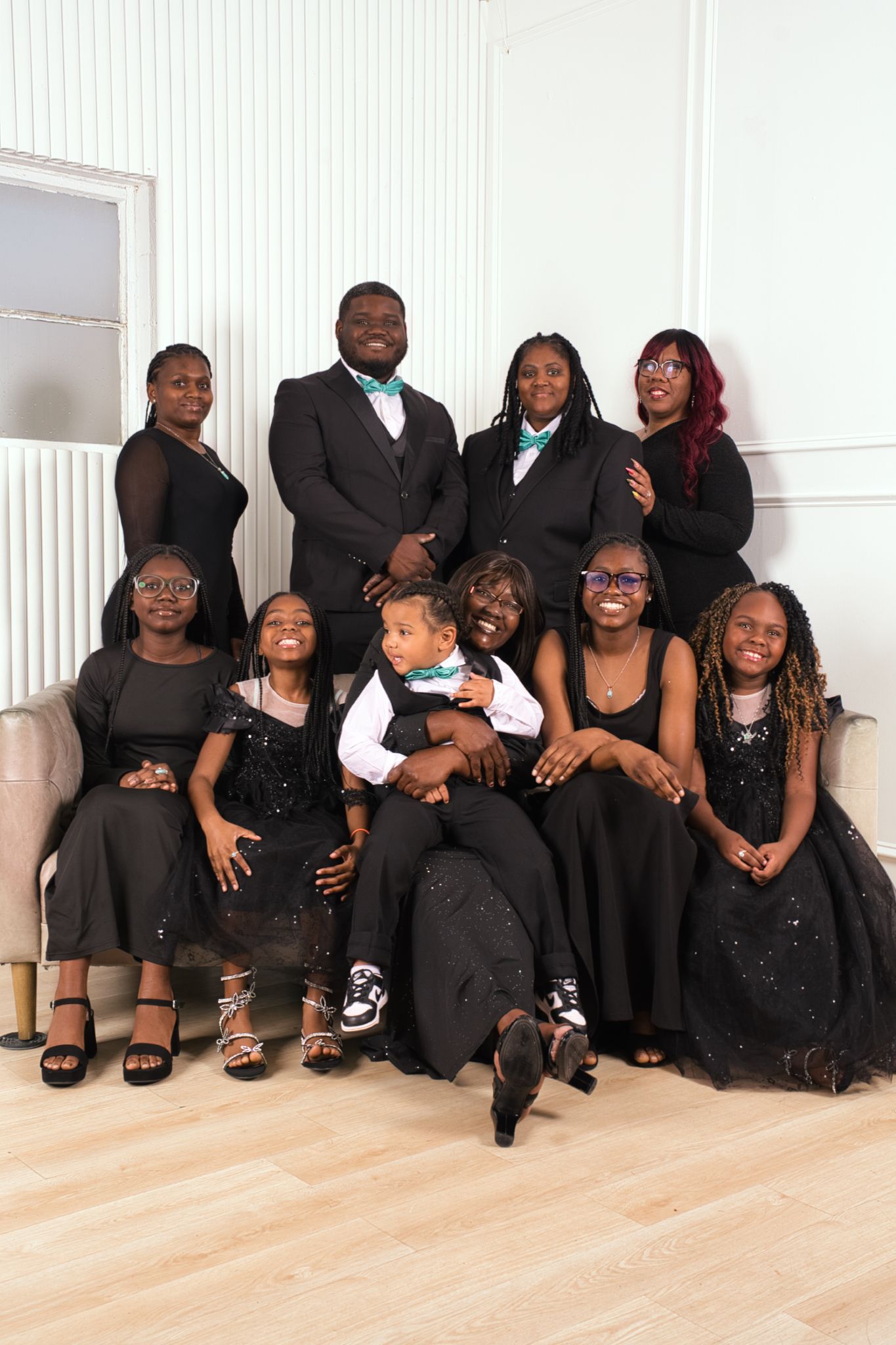 A large family group wearing coordinated black formal attire poses together sitting on a light wooden floor.