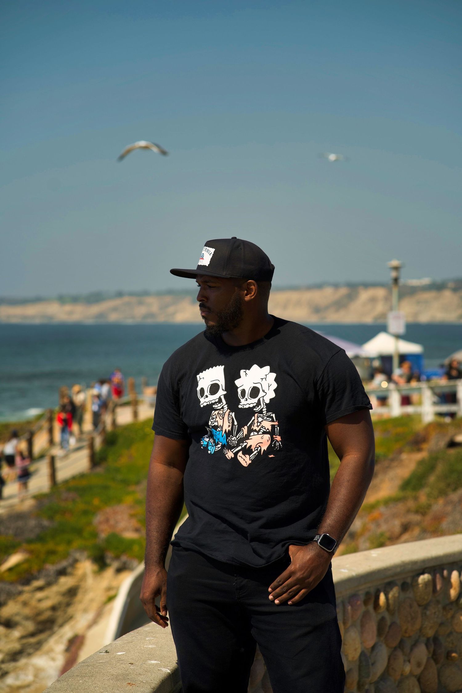 Person in black graphic t-shirt standing near coastal cliff with seagull flying overhead.