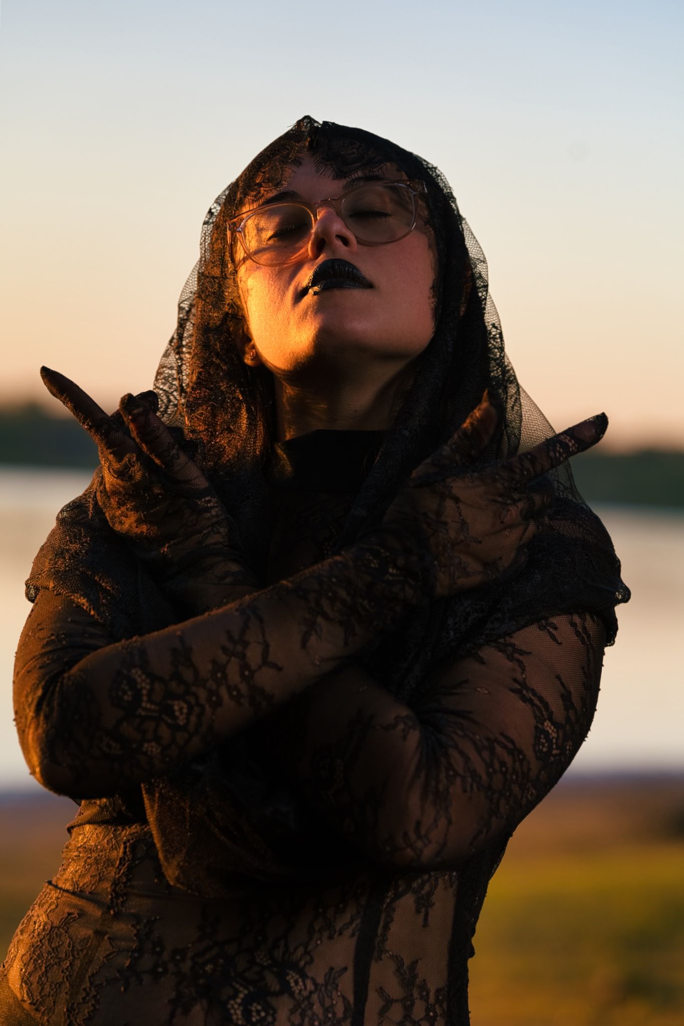 Figure in black lace veil poses dramatically against sunset sky.
