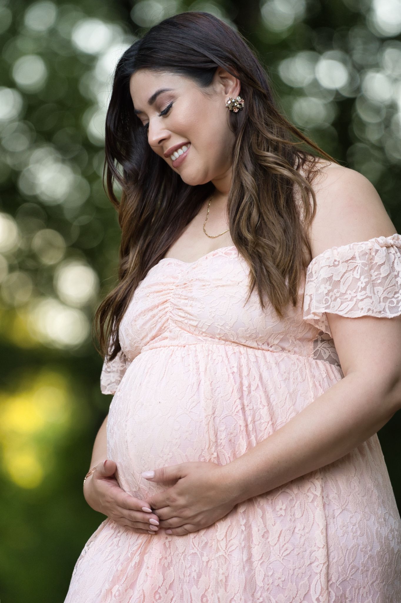Maternity portrait in pink lace dress with natural outdoor bokeh background.