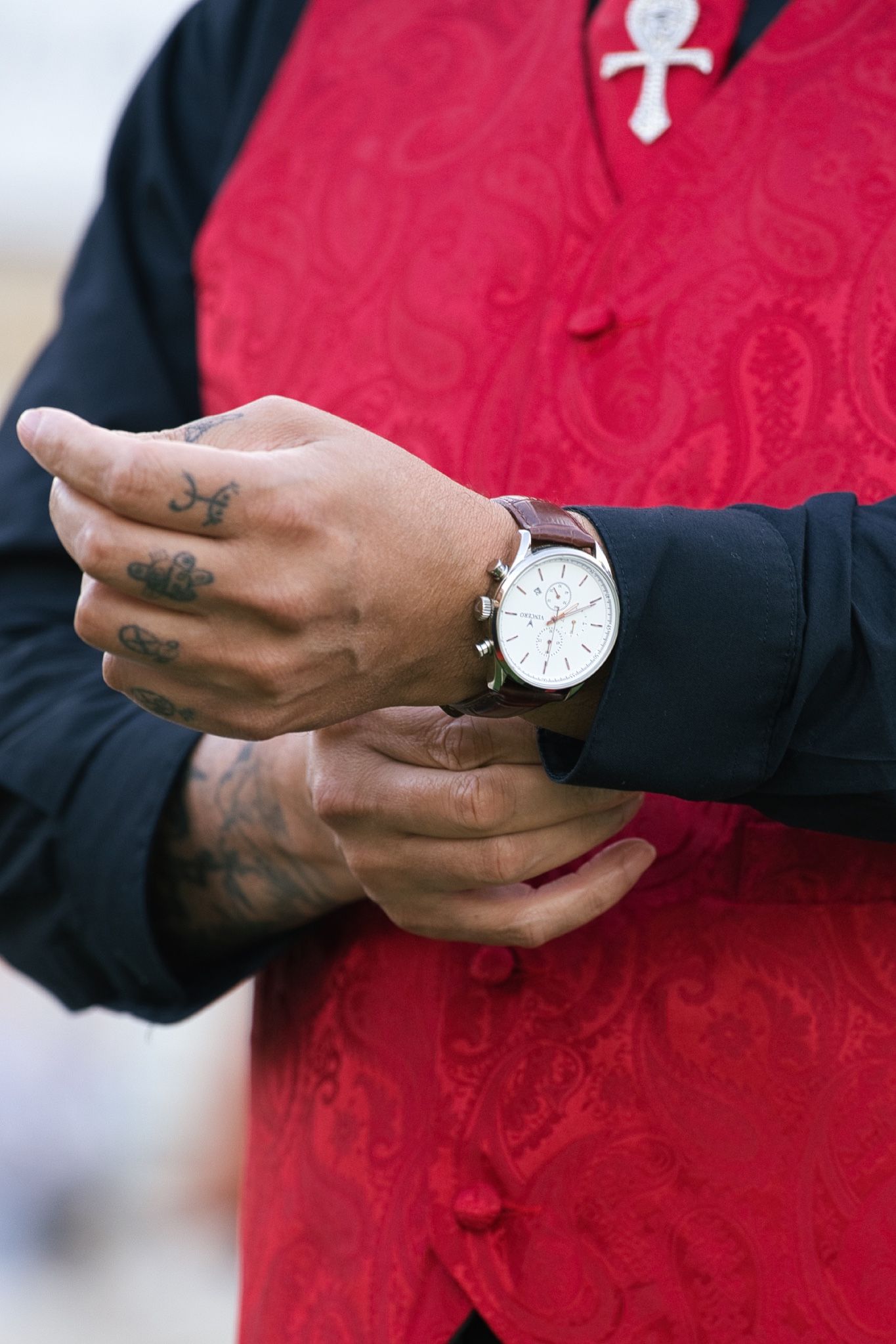 Close-up of hands with tattoos adjusting silver watch against red and black formal attire.