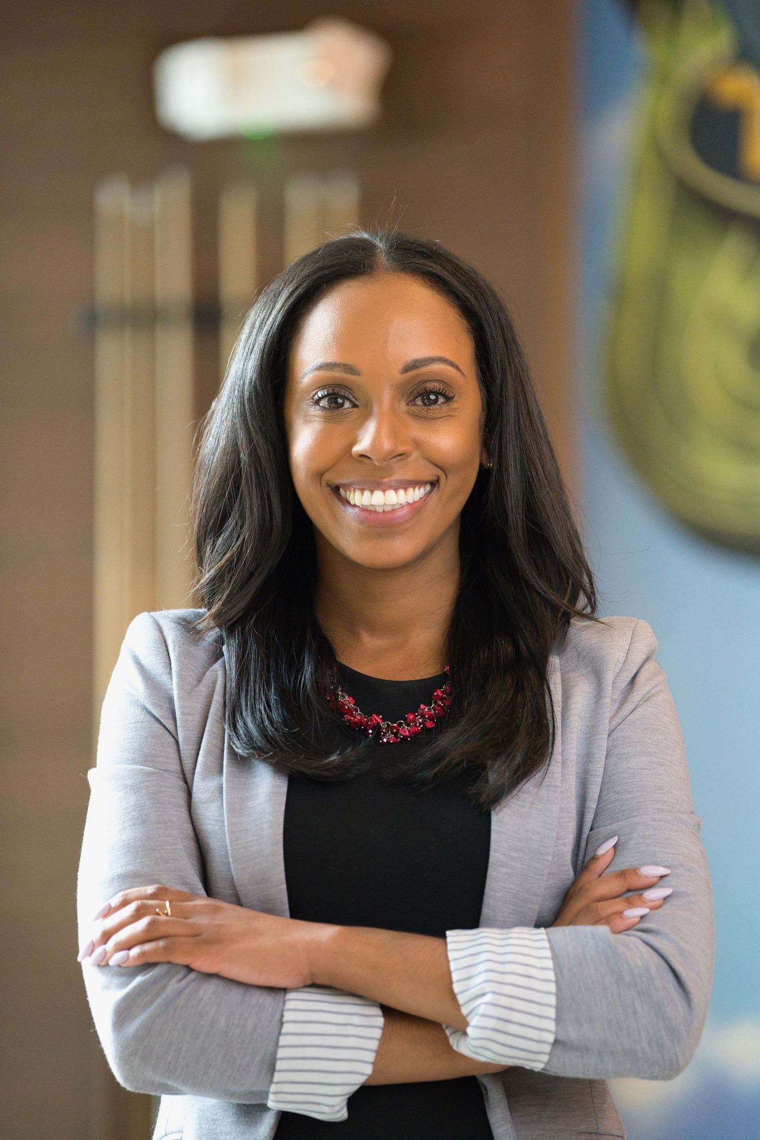Professional in a black top and gray blazer smiling warmly while standing with arms crossed in an office setting.