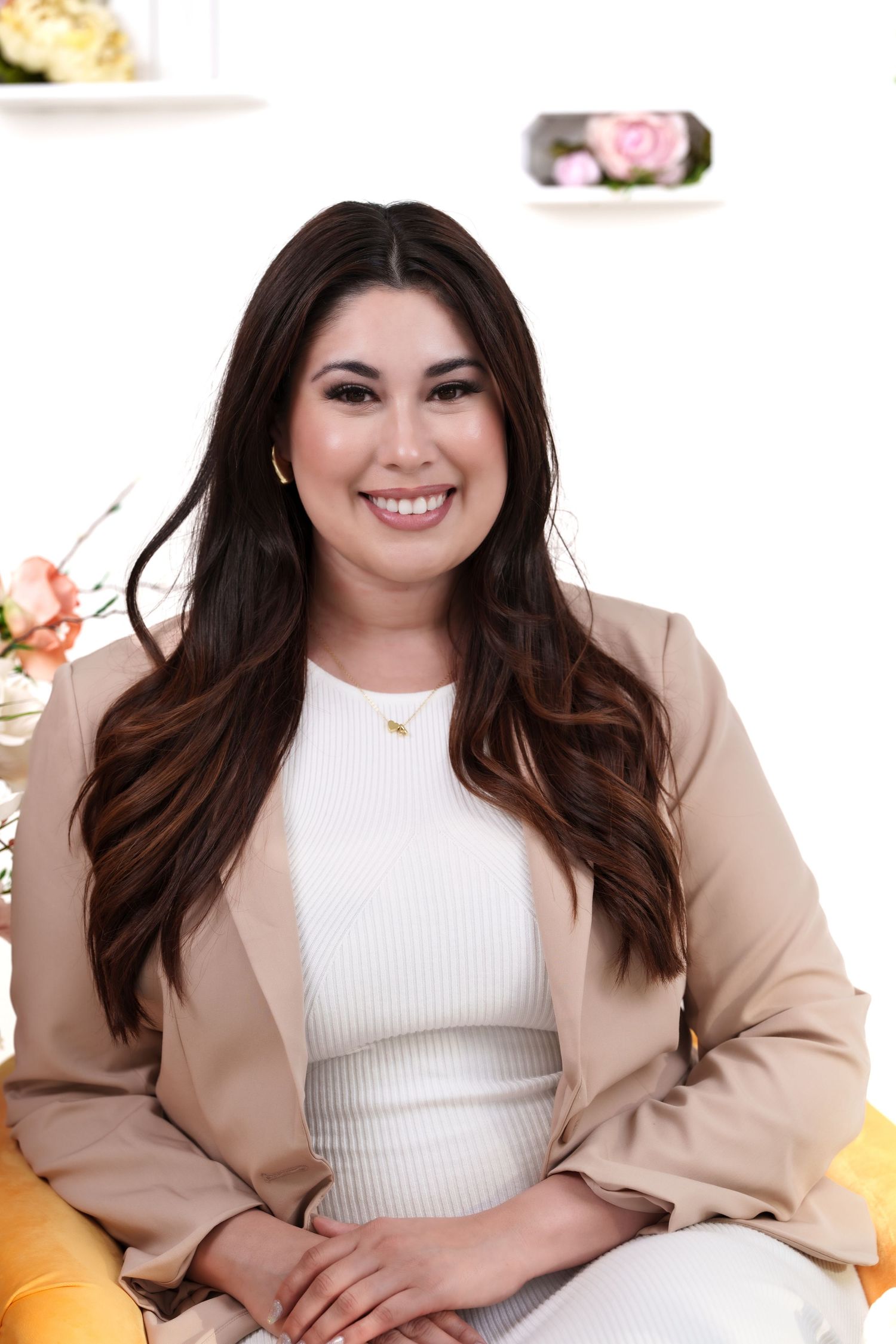 Portrait of someone in beige blazer sitting on yellow chair with floral background.