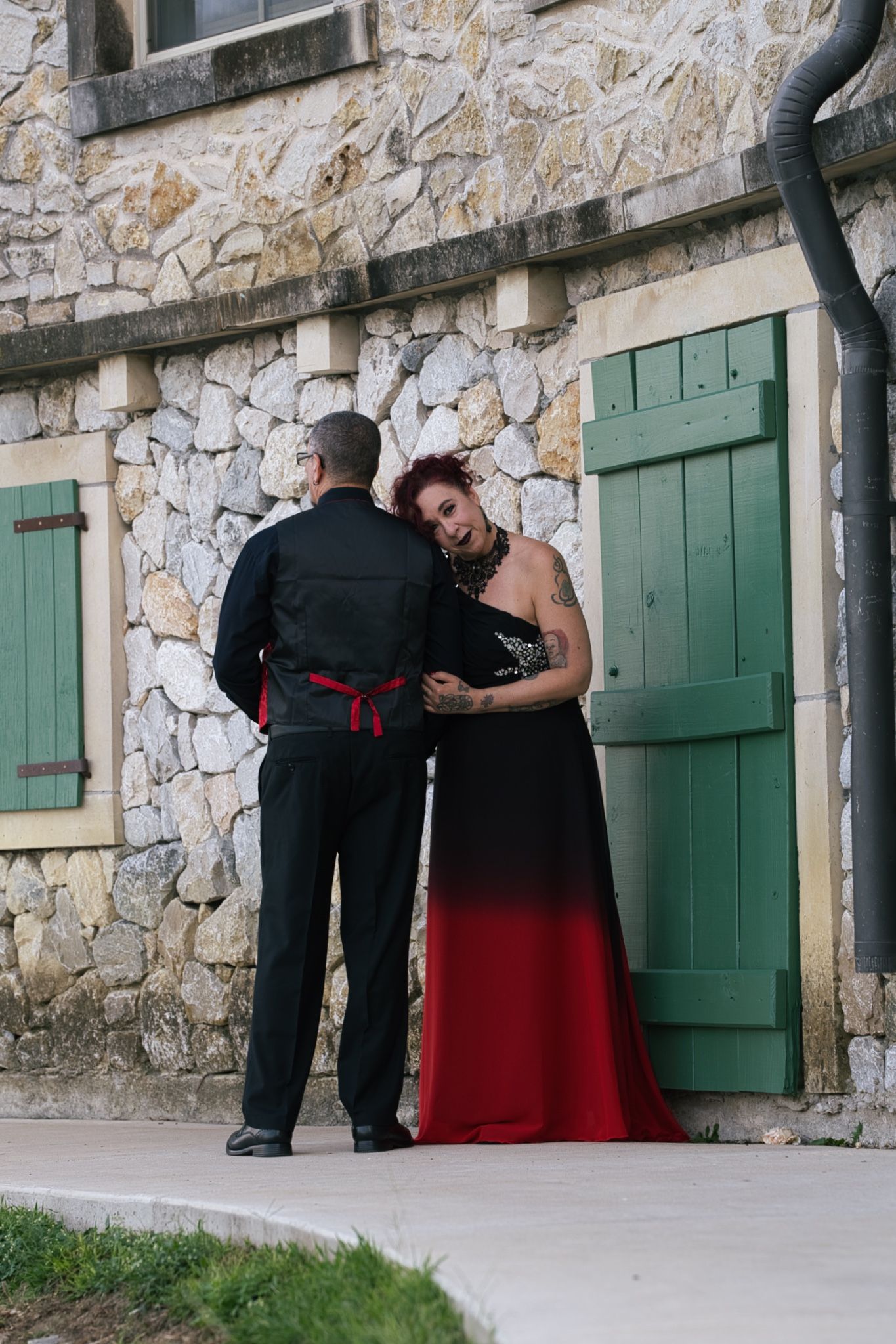 A couple in formal attire stands by stone wall with green shutters, groom in black and bride in red and black dress.