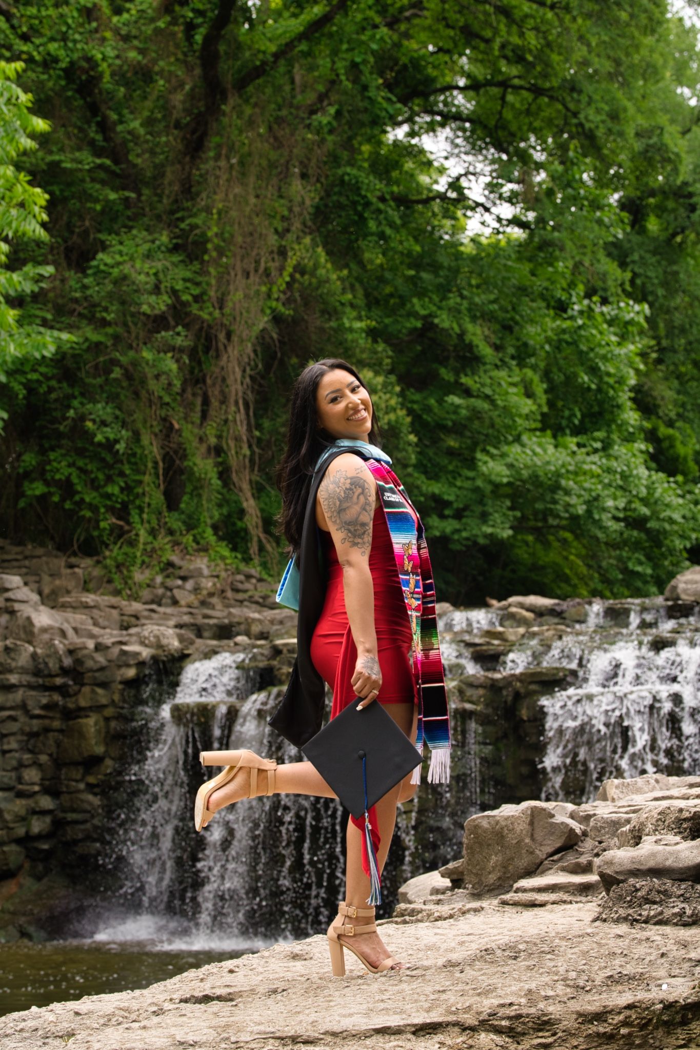 Graduate playfully poses with cap while standing on rocks near cascading waterfall.
