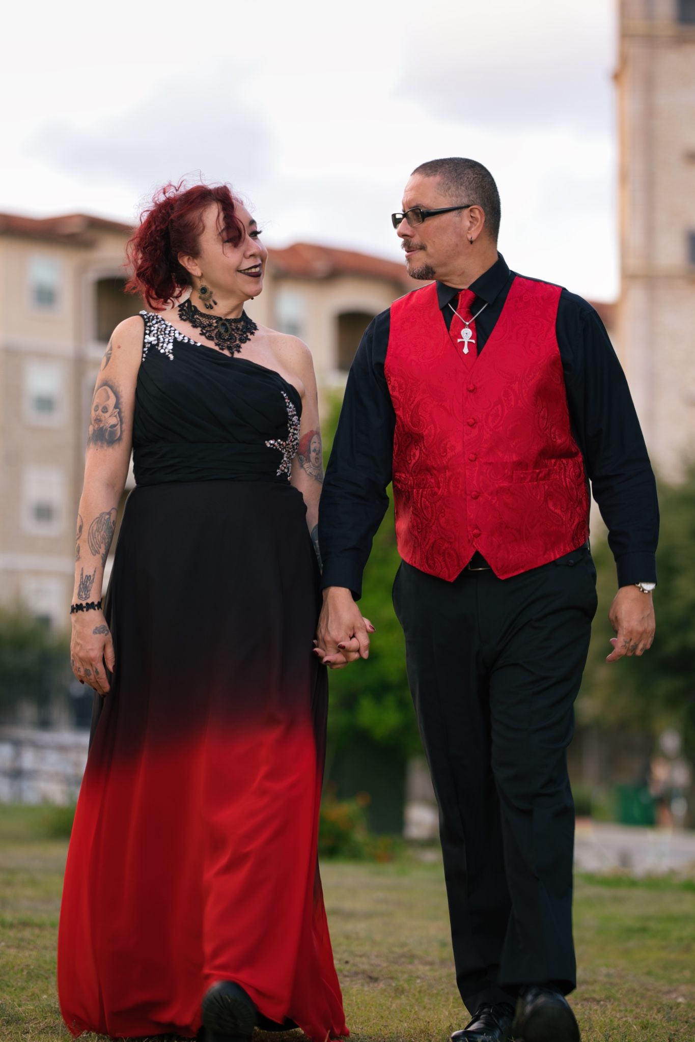 A couple walks hand in hand outdoors, wearing coordinated formal red and black attire.