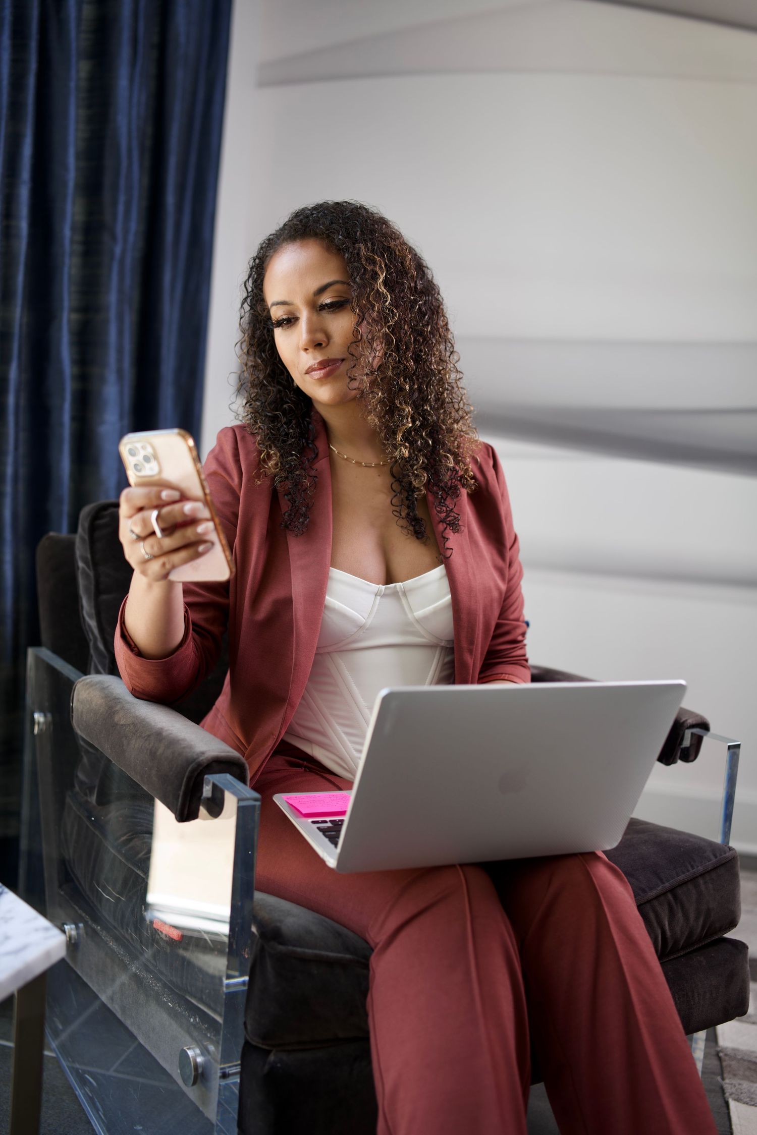 Professional woman in burgundy suit works on laptop and checks phone while seated.