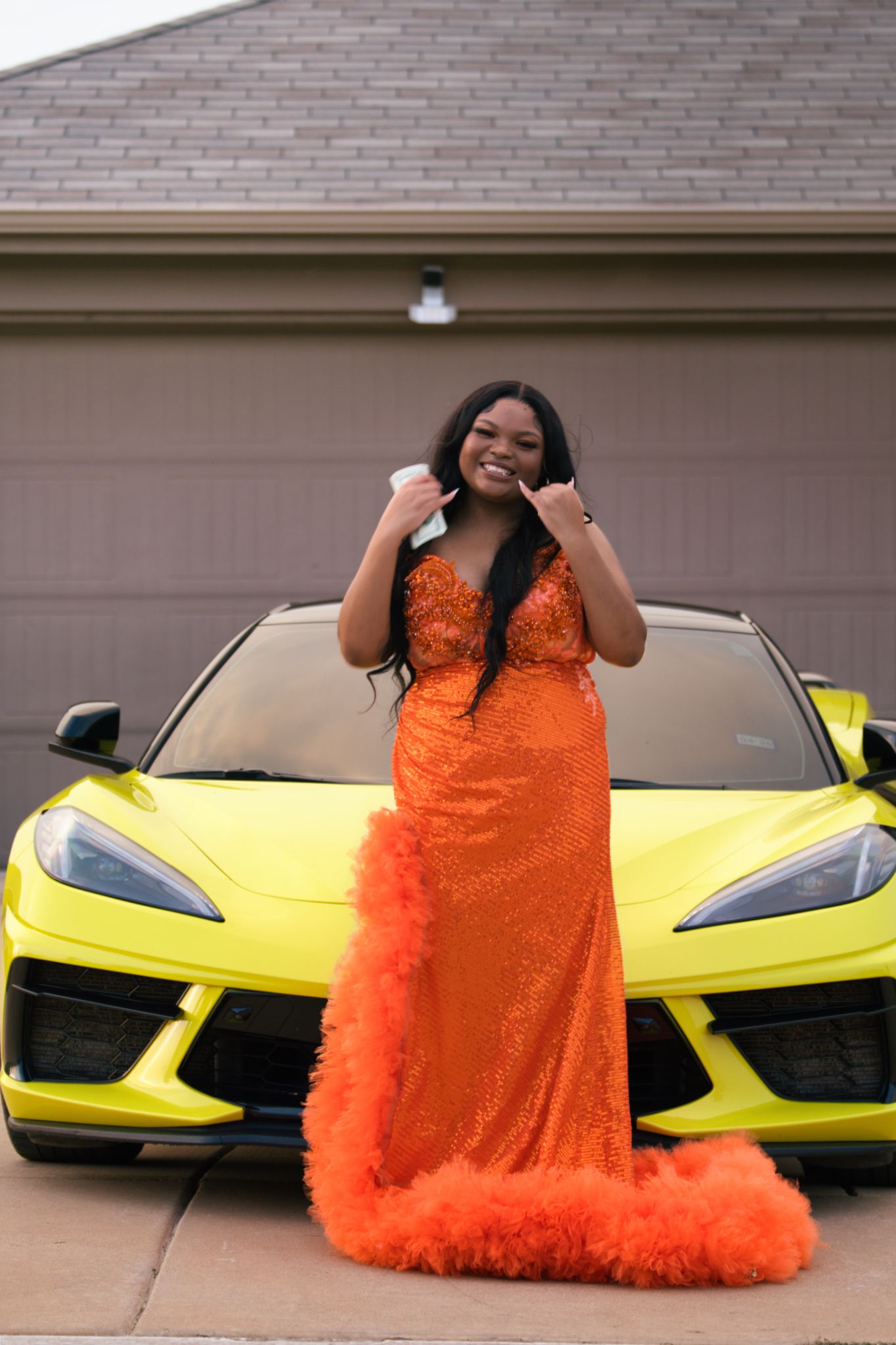 Young woman in orange sequin gown with feather trim posing in front of yellow Corvette sports car.