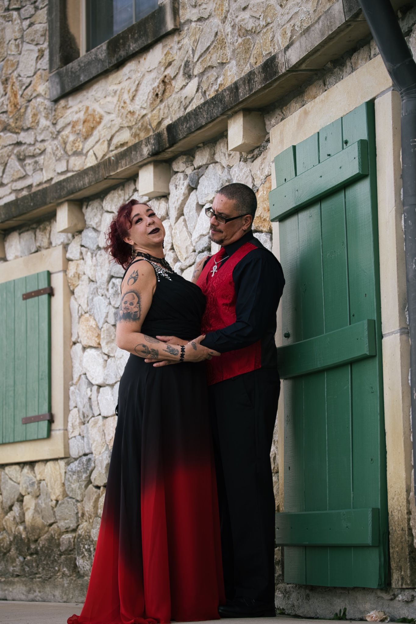A couple in formal attire embraces against a rustic stone wall with green shutters, wearing black and red coordinated outfits.