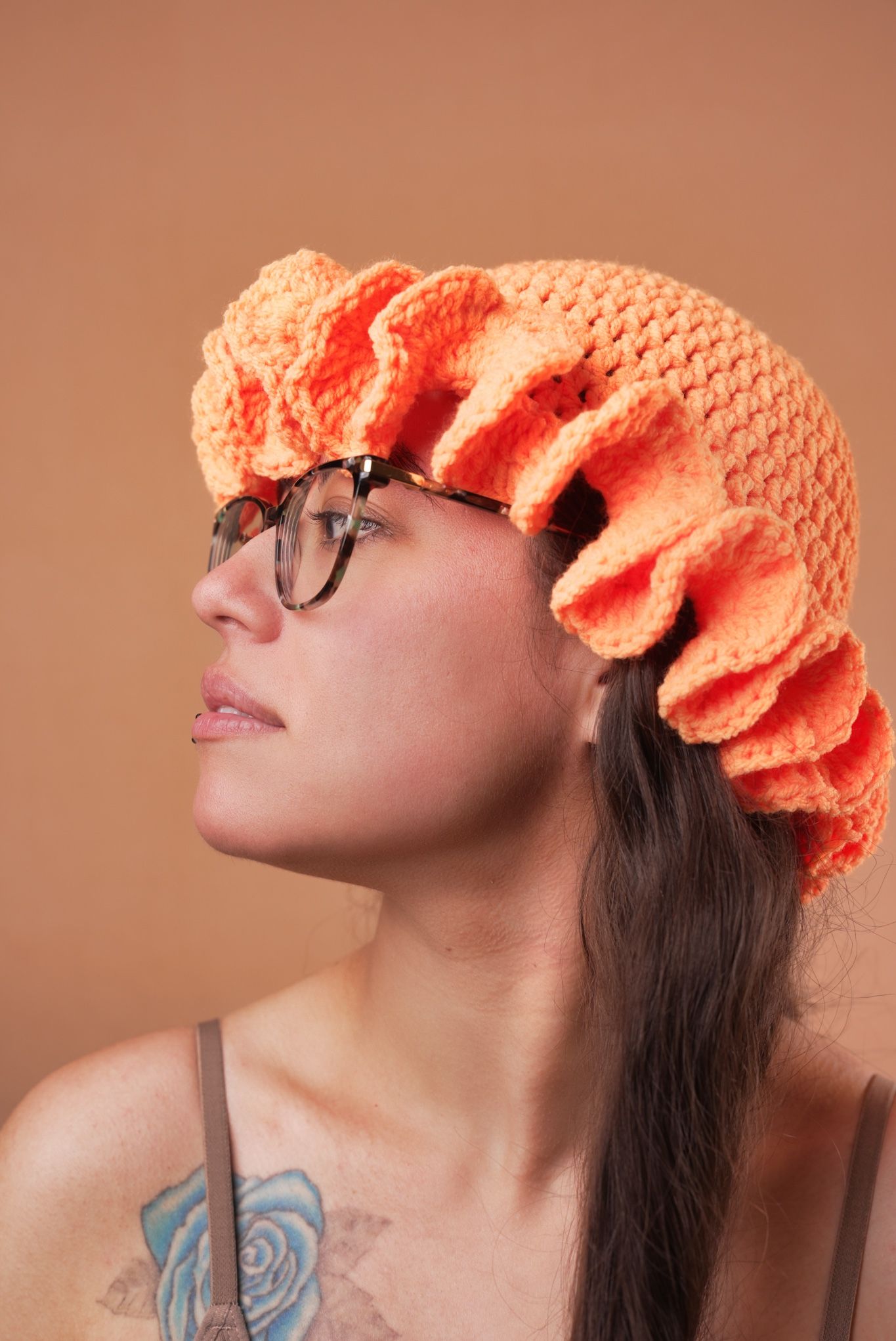 Side profile portrait of person wearing glasses and coral knit hat against peach background.