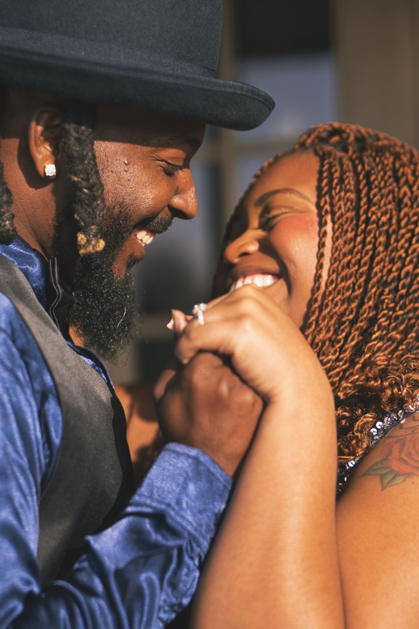 A joyful Black couple laughing and holding hands, the man wearing a black hat and blue shirt, the woman with braided hair.