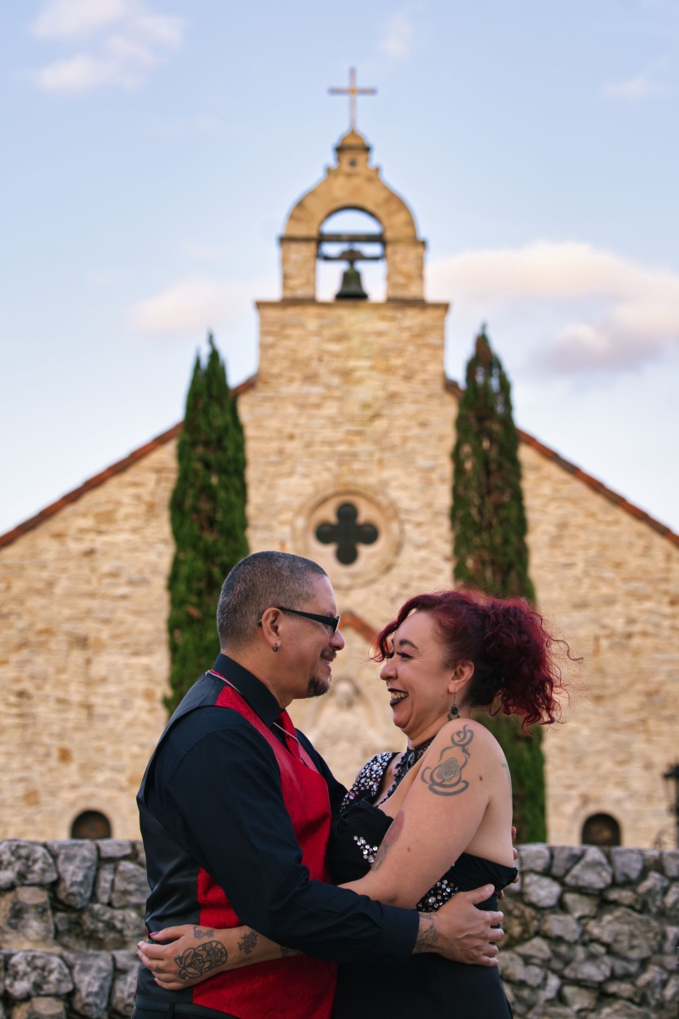 A couple embraces and laughs in front of a stone church with cypress trees at sunset.