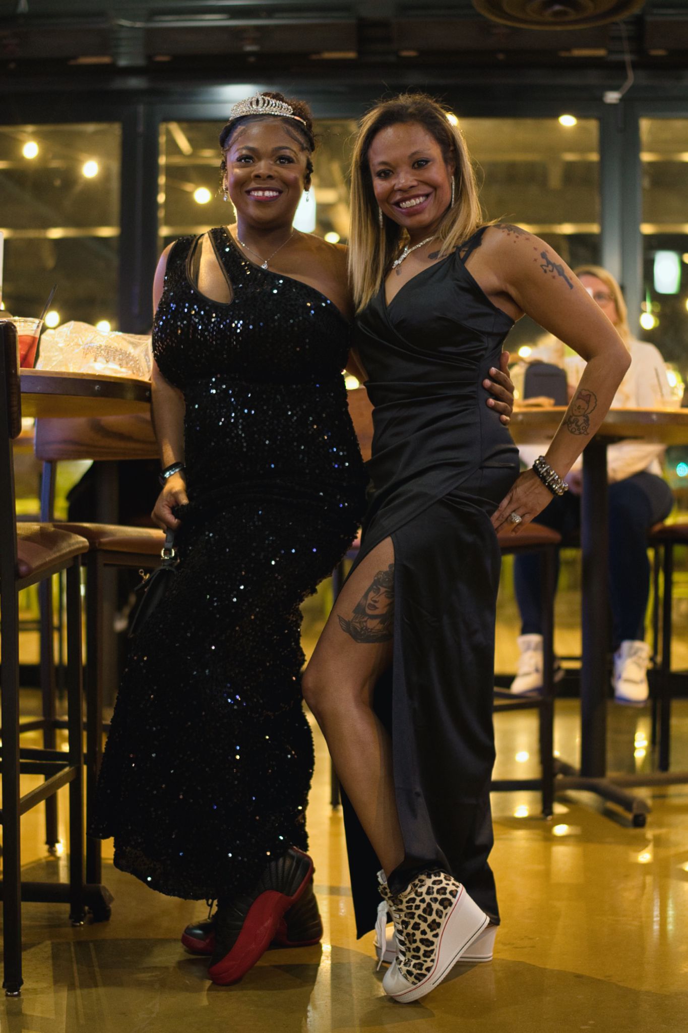 Two people in black evening attire pose playfully in a dimly lit venue with warm ambient lighting.