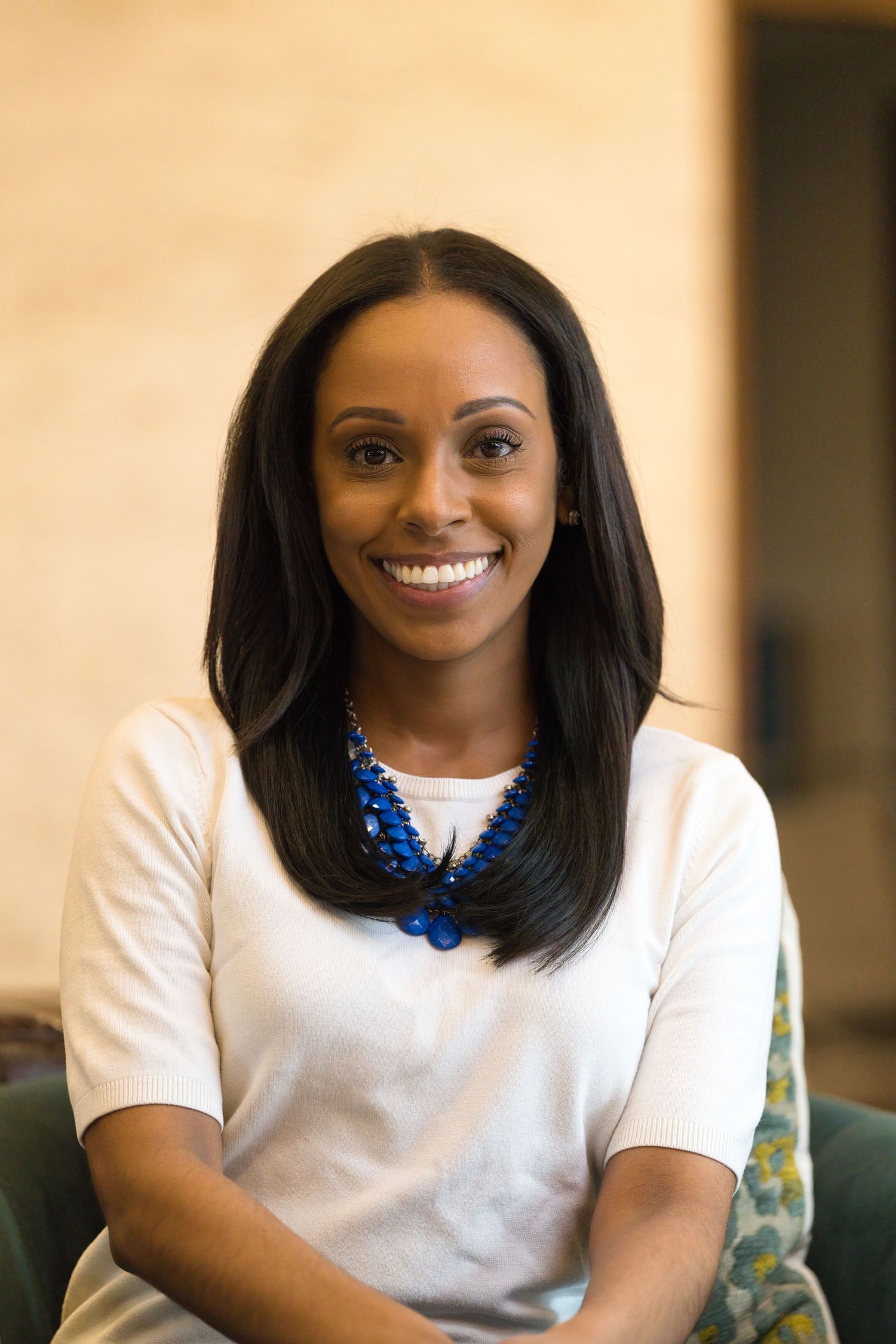 A person wearing a white t-shirt and blue statement necklace sits on a couch with a warm-toned background.