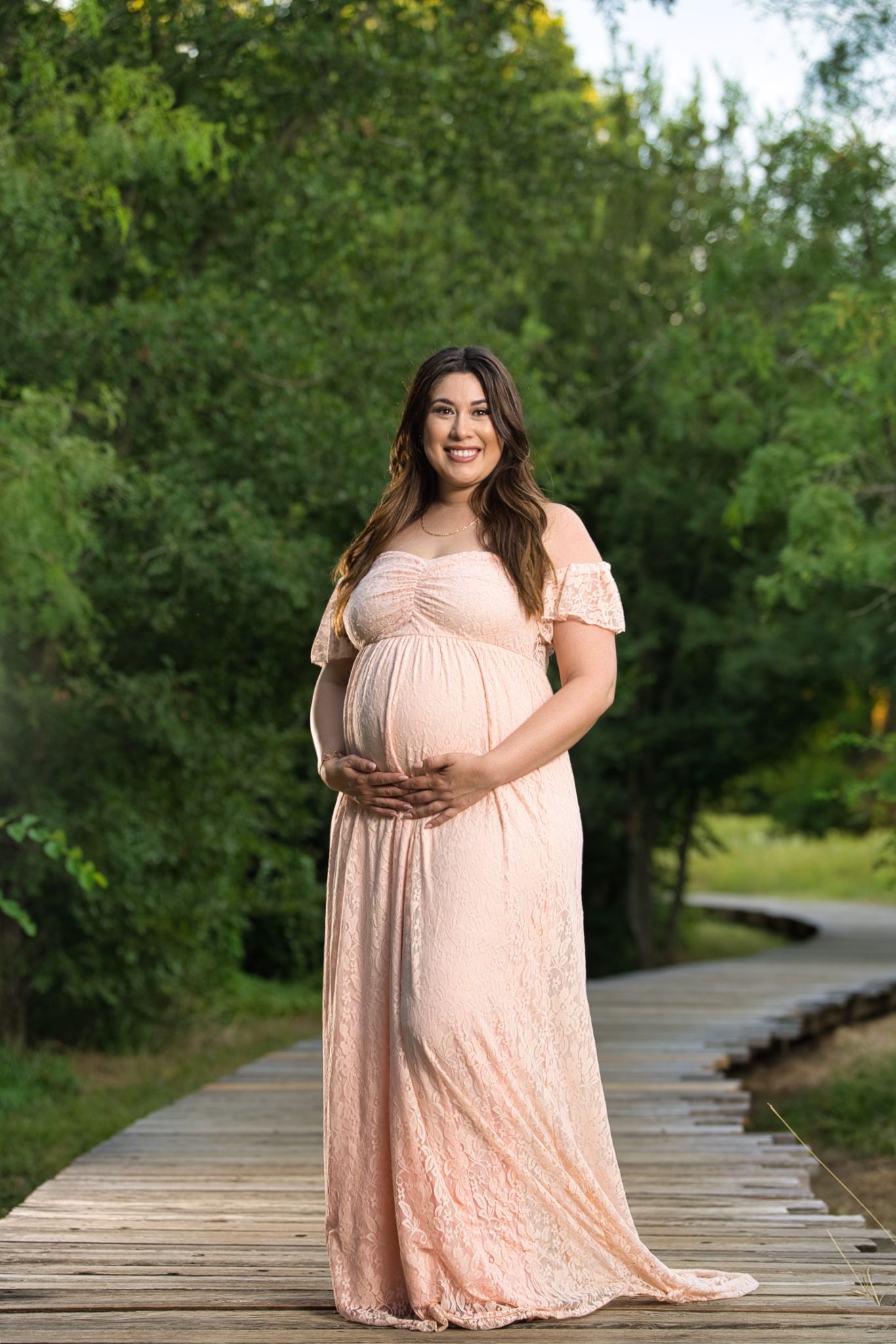 Maternity photo of woman in blush gown standing on wooden bridge surrounded by greenery.