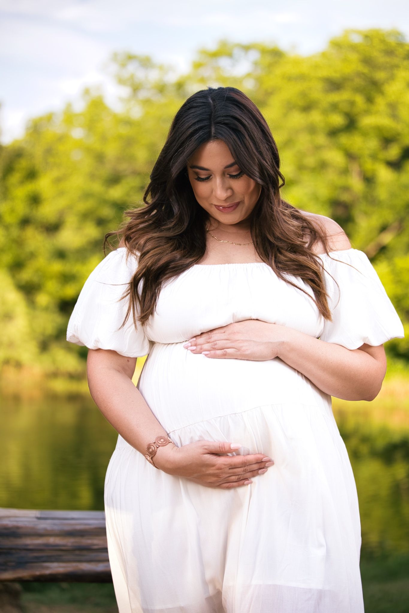 Expectant mother in white dress poses by lake during pregnancy photoshoot.