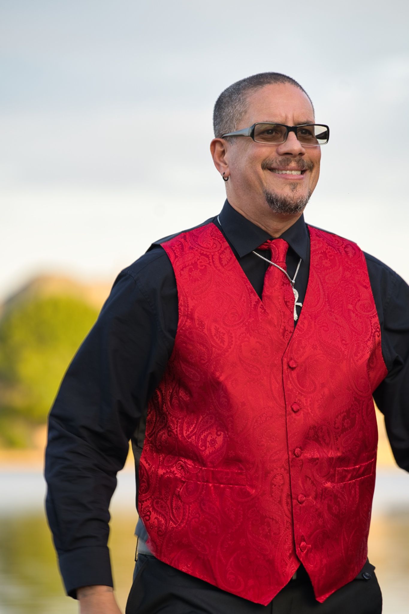 A person wearing glasses and a red brocade vest with black shirt and tie smiles outdoors.