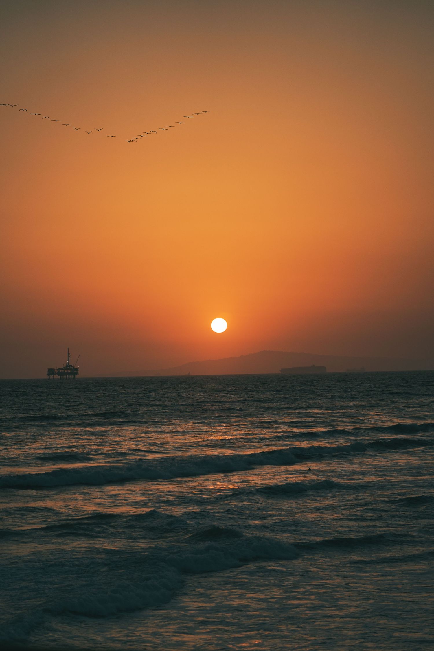 Golden sunset over ocean with birds flying and oil rig silhouetted on horizon.