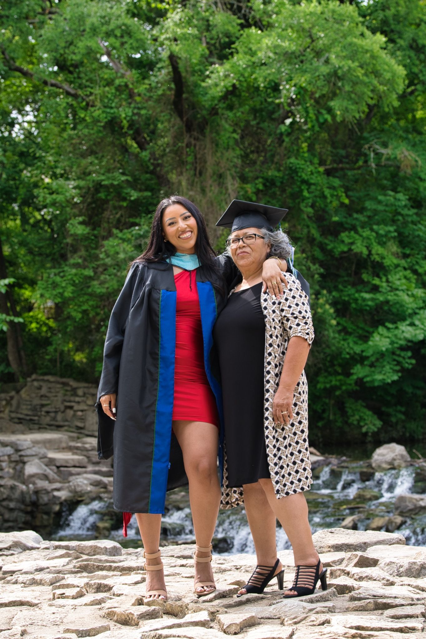 Graduate in cap and gown poses with family member on rocky terrain by waterfall.